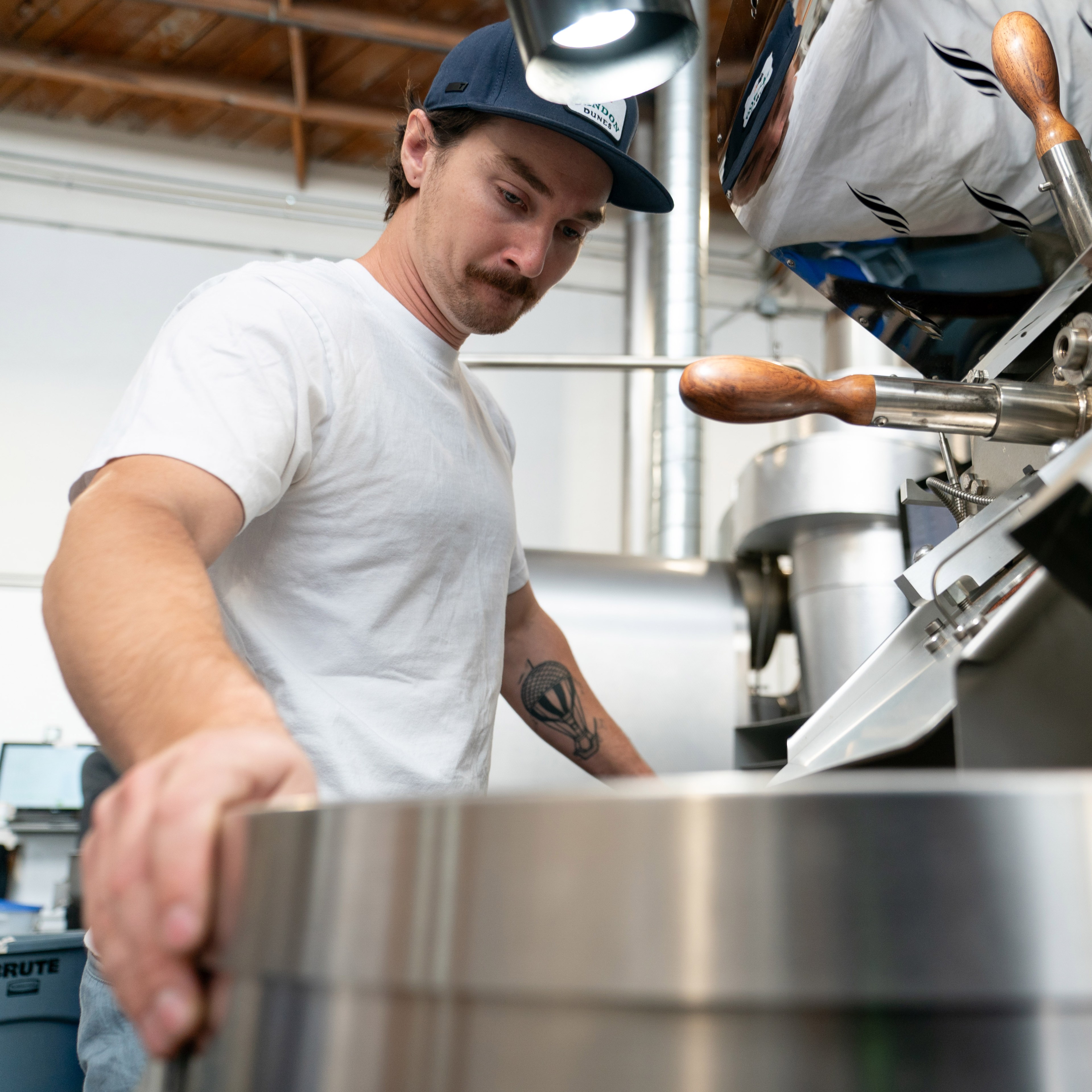 A man in a white shirt and blue cap operates a large coffee roaster in an industrial setting. He has a focused expression and a hot air balloon tattoo on his arm.