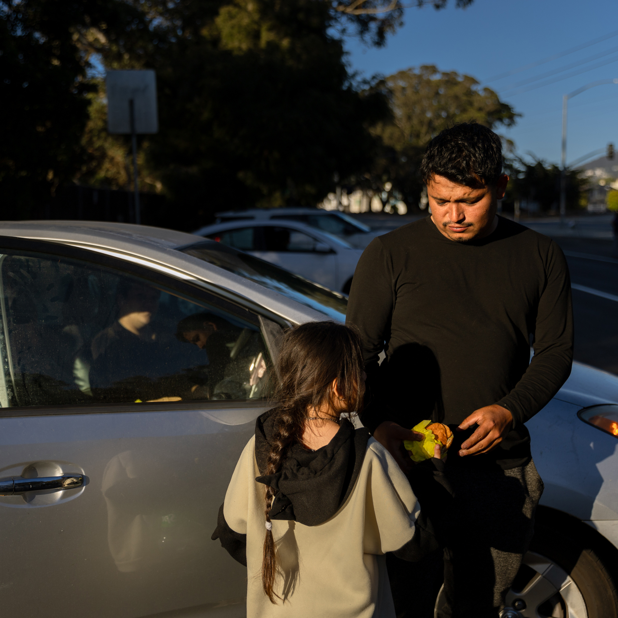 A man in a black shirt stands by a car holding food, while a girl with a long braid, wearing a hoodie, looks up at him. Several cars and trees are in the background.