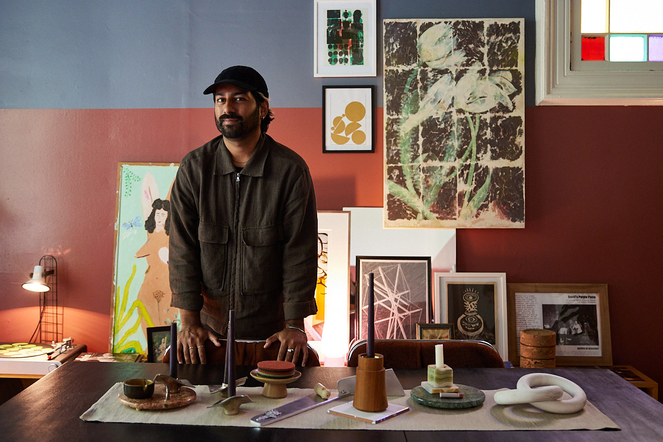 A man stands behind a table with art and candles in a room decorated with various paintings and framed pictures. A stained glass window is on the right.