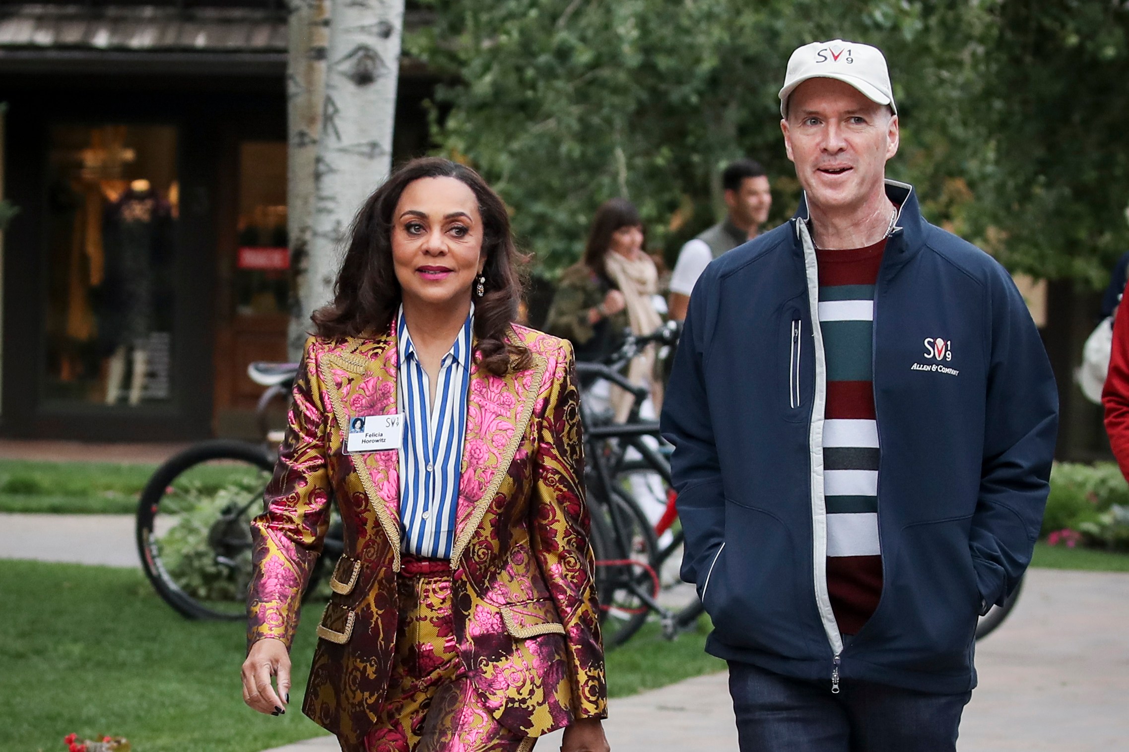 A woman in a vibrant, pink-and-gold baroque-style suit walks alongside a man in casual attire with a striped shirt and a head cap on a paved path with greenery.