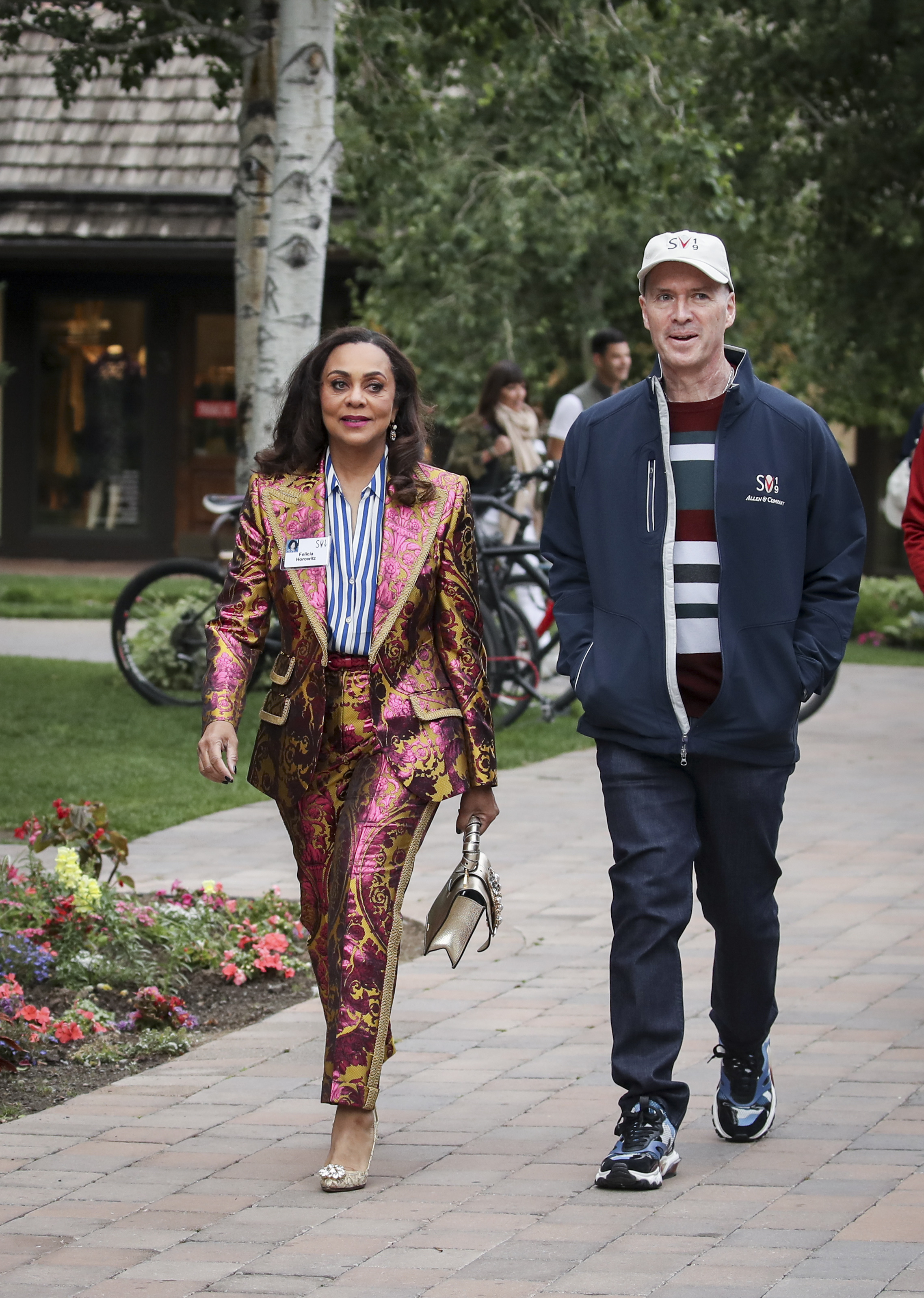 A woman in a vibrant, pink-and-gold baroque-style suit walks alongside a man in casual attire with a striped shirt and a head cap on a paved path with greenery.