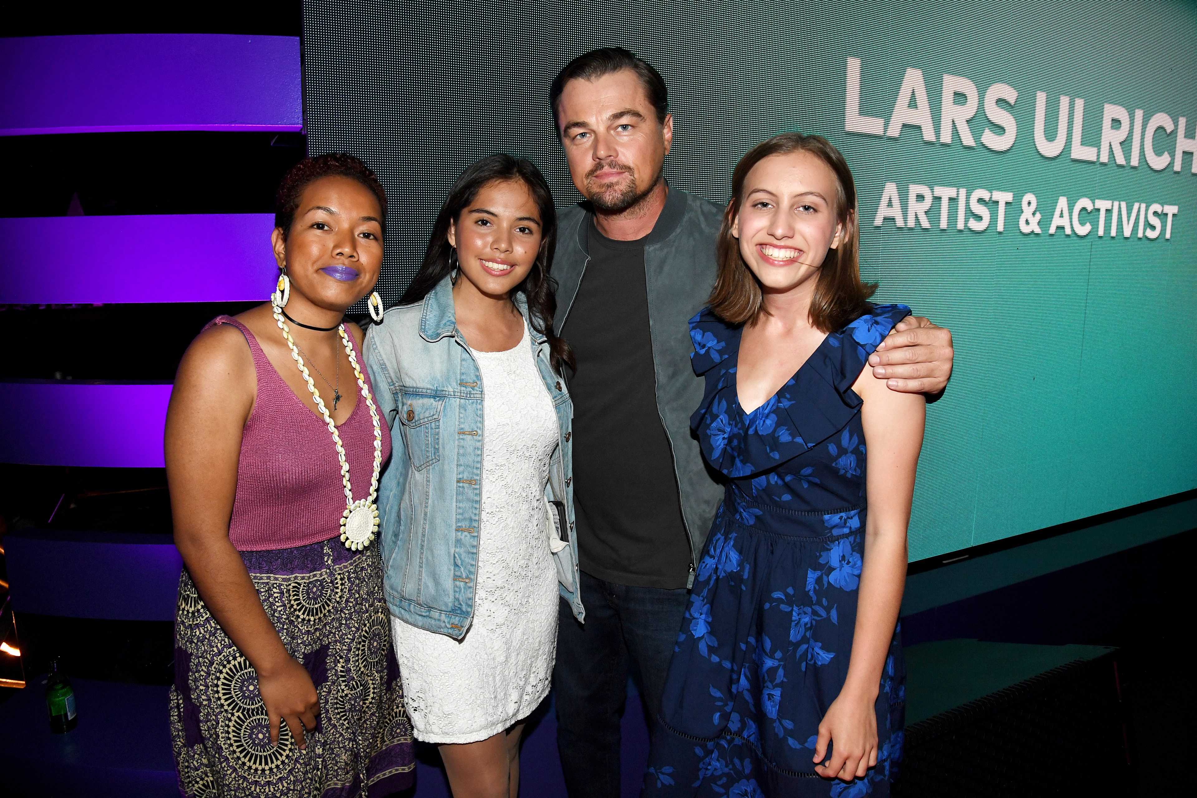 A man and three women smile for a photo; the man wears a black jacket and the women wear colorful dresses. The background features the text &quot;Lars Ulrich, Artist &amp; Activist.&quot;