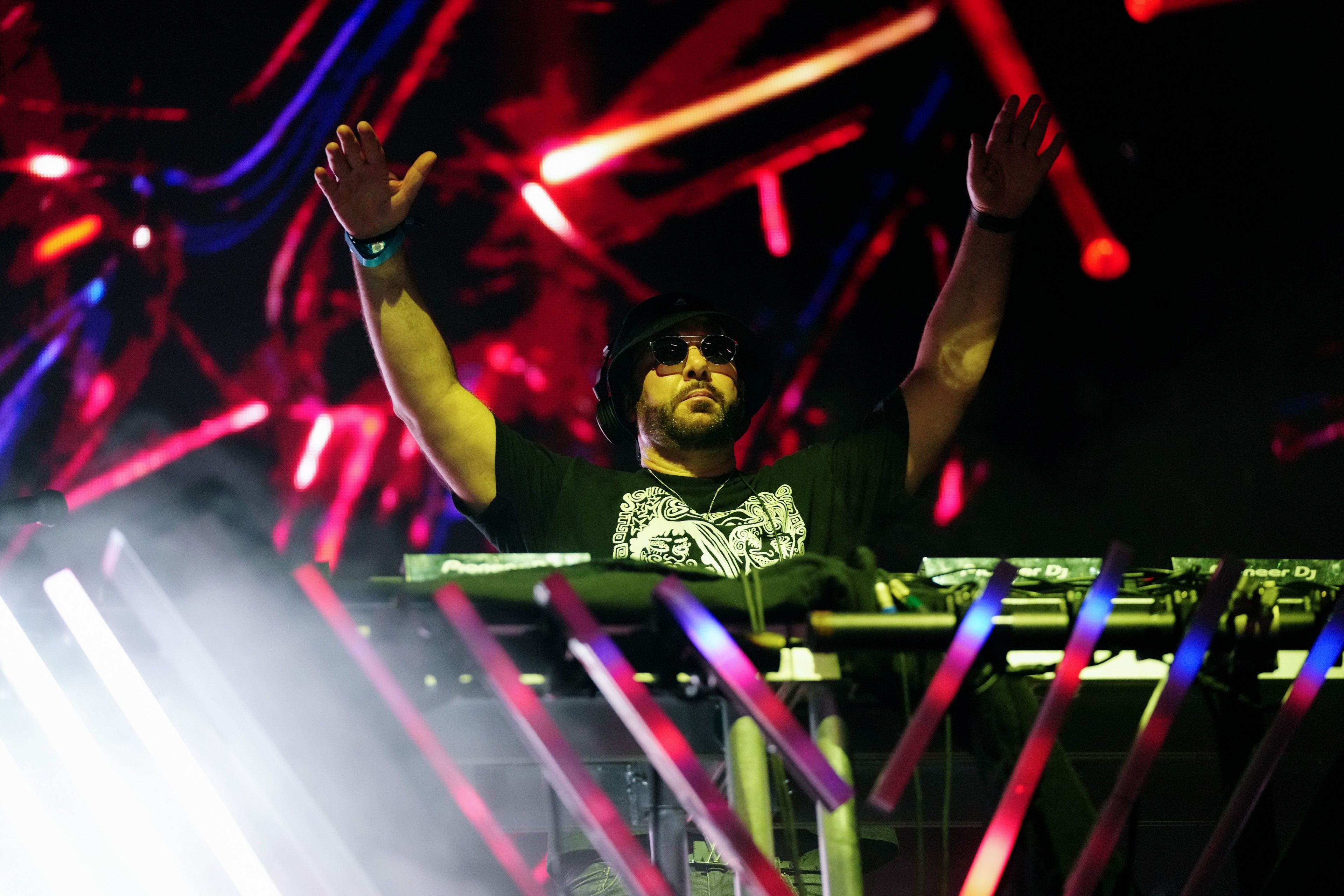 A DJ wearing sunglasses and a black hat stands behind mixing equipment with both hands raised. The background features vibrant red, blue, and purple lights.