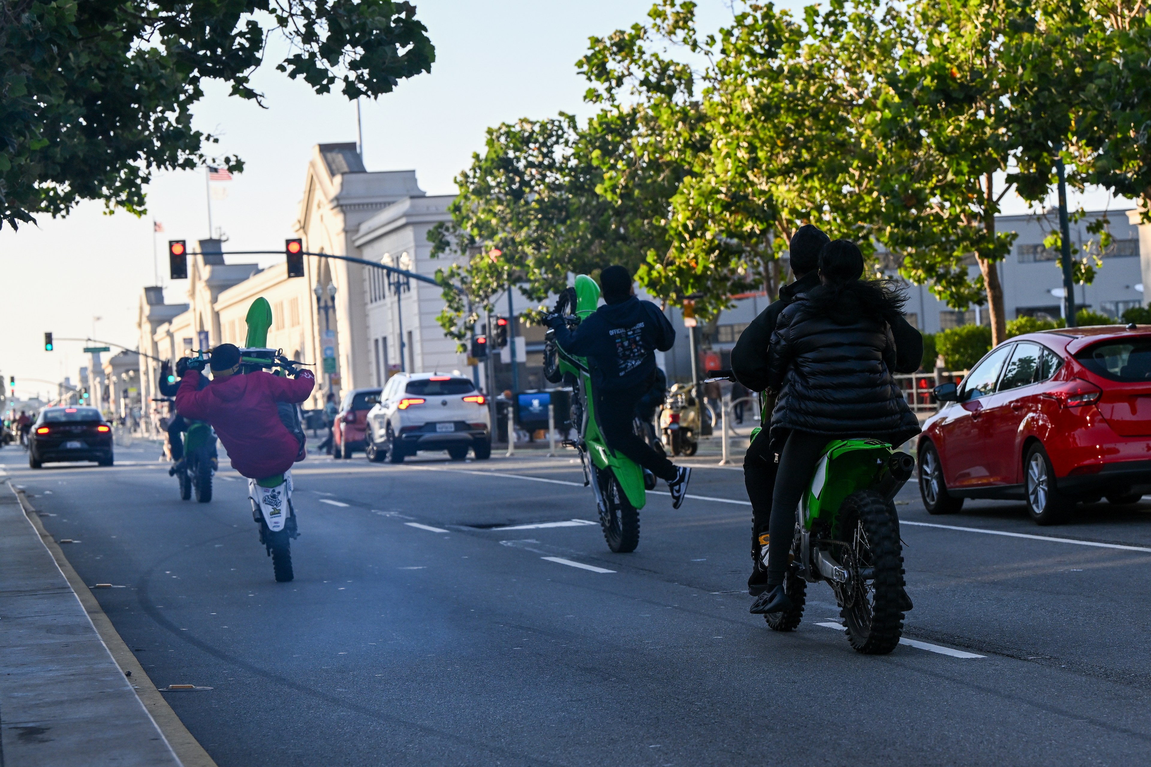 People are performing wheelies on dirt bikes on a city street, surrounded by cars and trees, with buildings and traffic signals visible in the background.