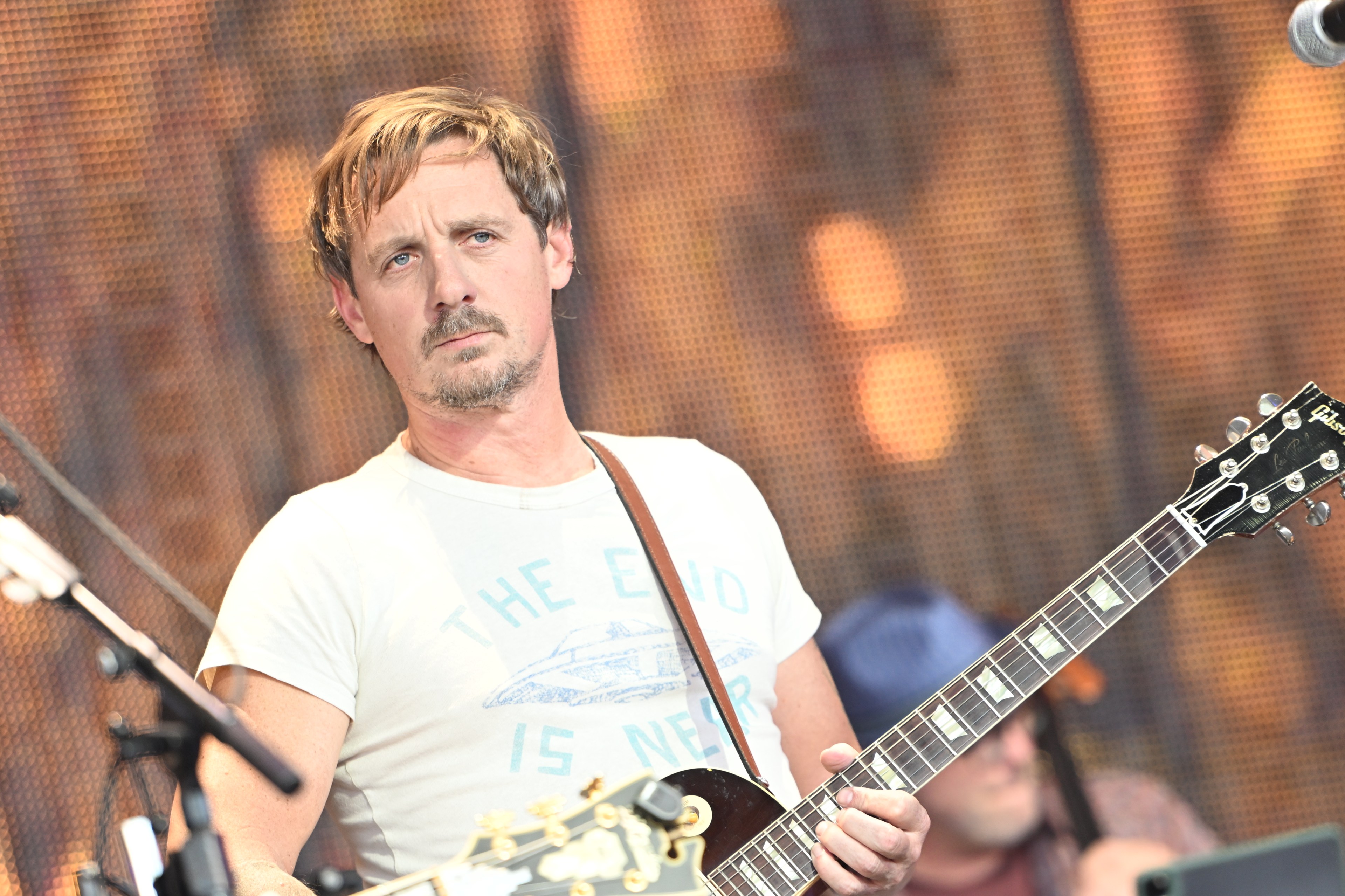 A man with short, light brown hair and a mustache plays an electric guitar. He wears a T-shirt with &quot;THE END IS NEAR&quot; printed on it, against a blurred, colorful background.