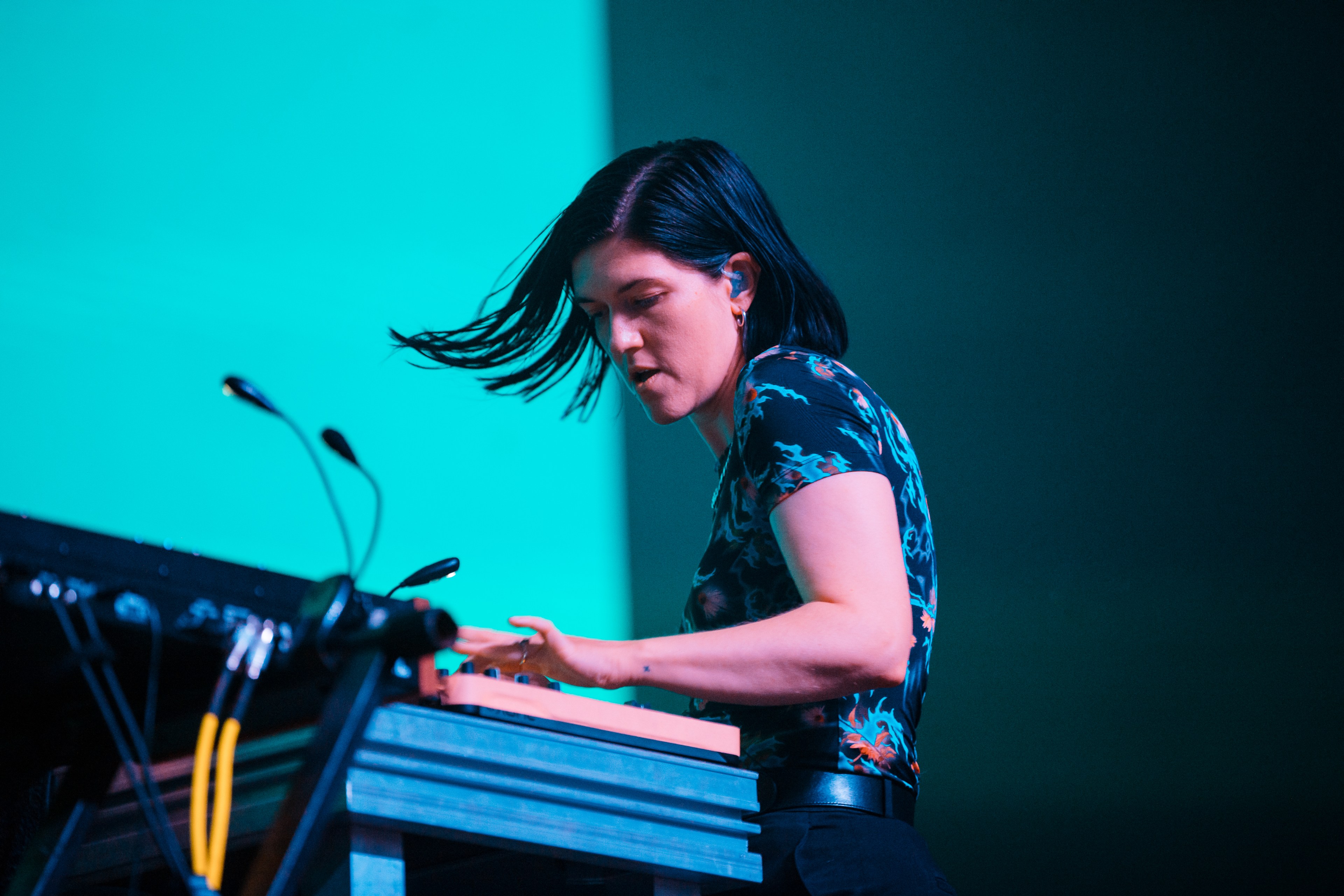 A person with short black hair is playing an electronic keyboard on stage, dressed in a patterned shirt against a blue background, focused intently on the performance.