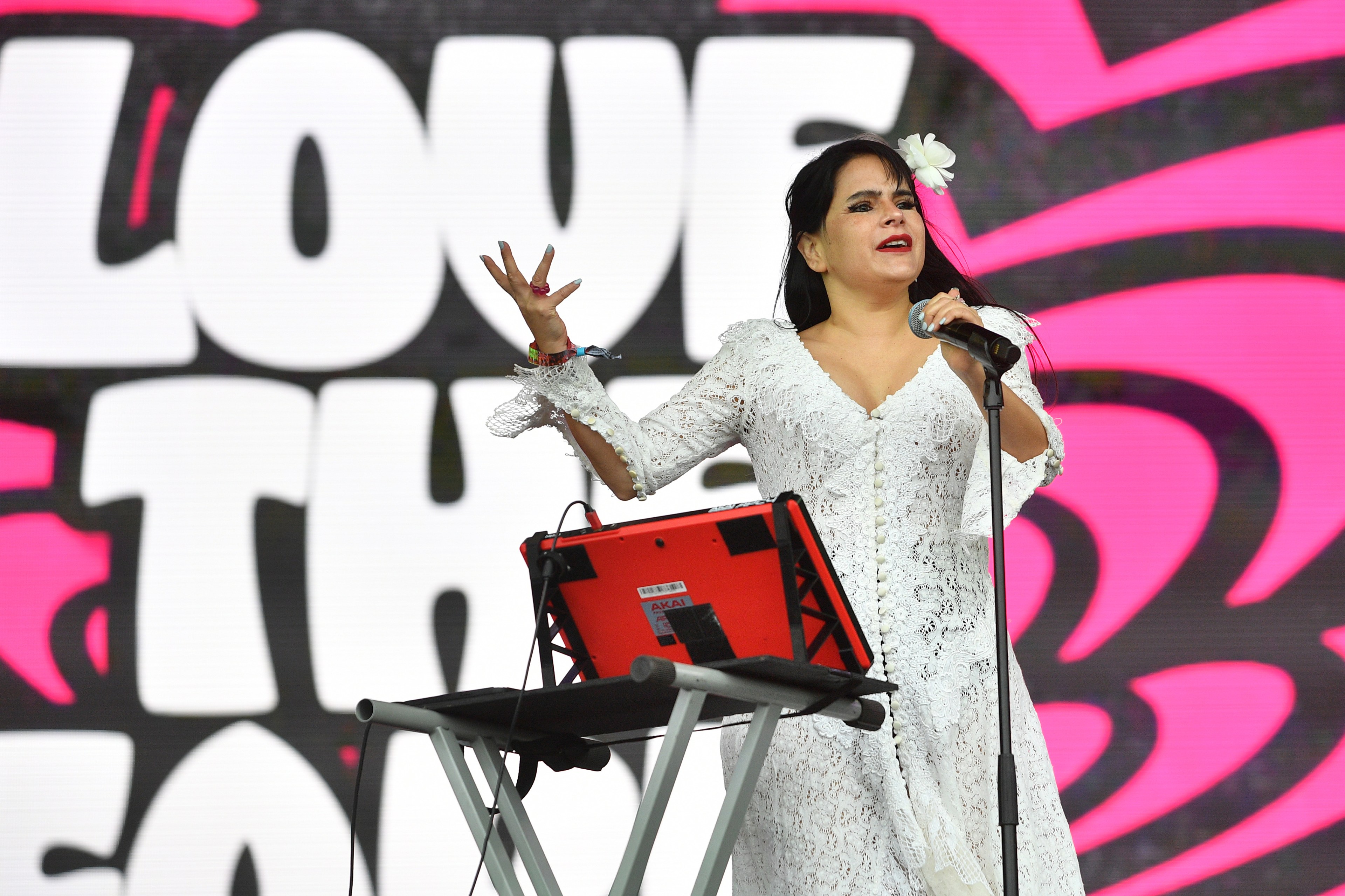 A woman in a white lace dress with a flower in her hair sings passionately with a microphone on stage, with a colorful &quot;Love&quot; sign in the background.