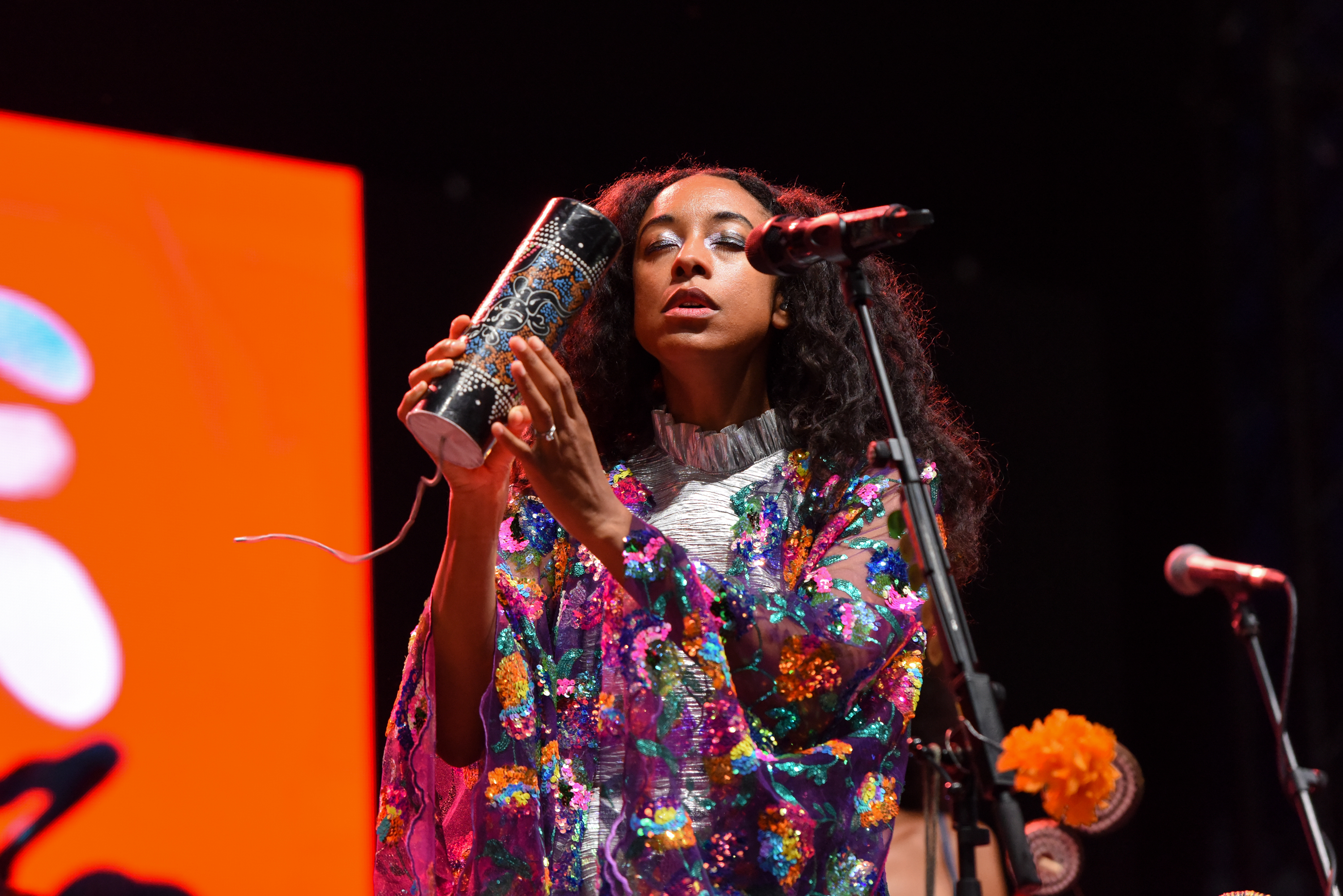 A woman in a colorful, floral-patterned outfit is performing on stage, holding a decorated cylindrical instrument near a microphone with a red and black background.