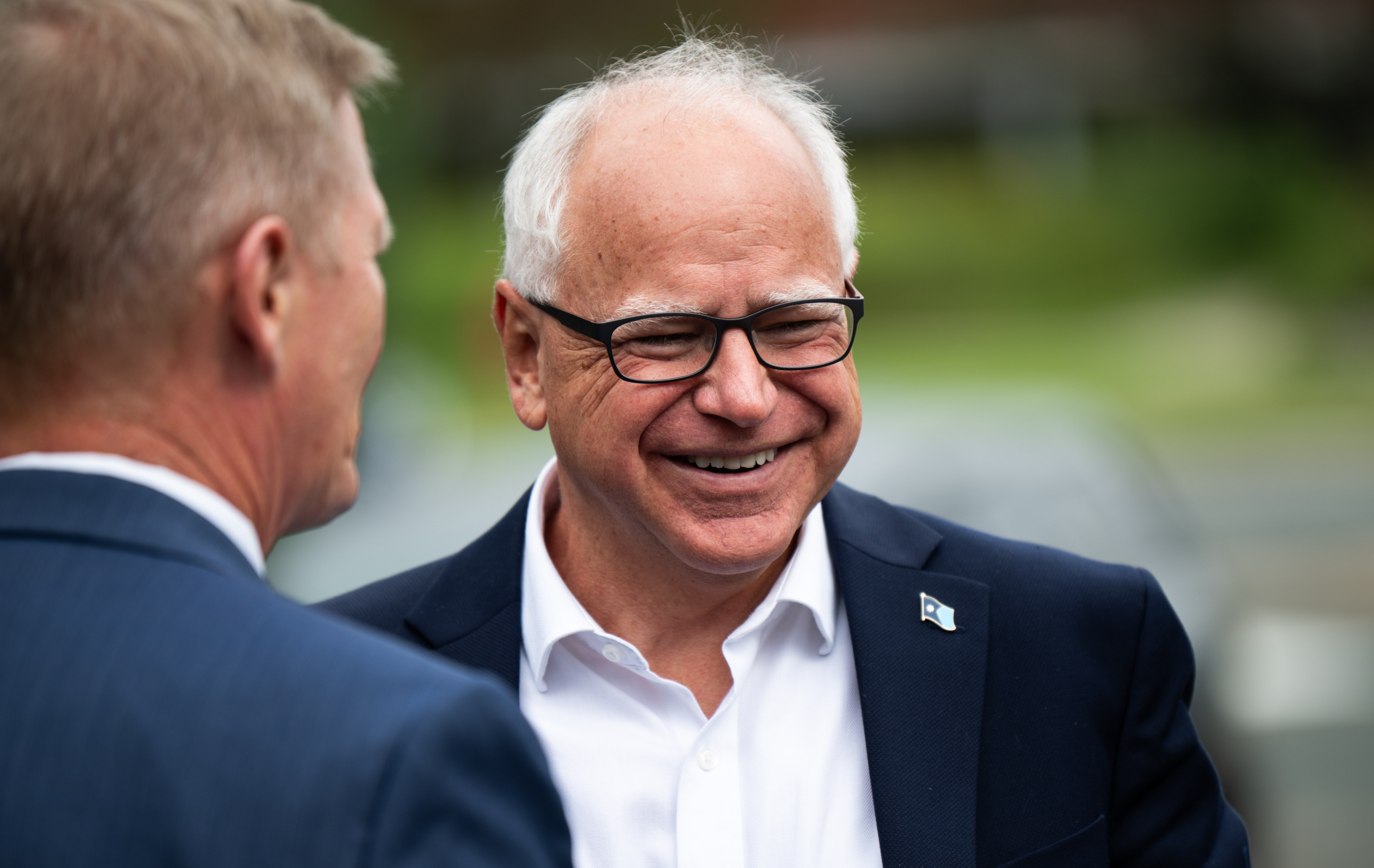 Two men in suits are outdoors, one with white hair and glasses smiling warmly, engaging in conversation, with a blurred green background.