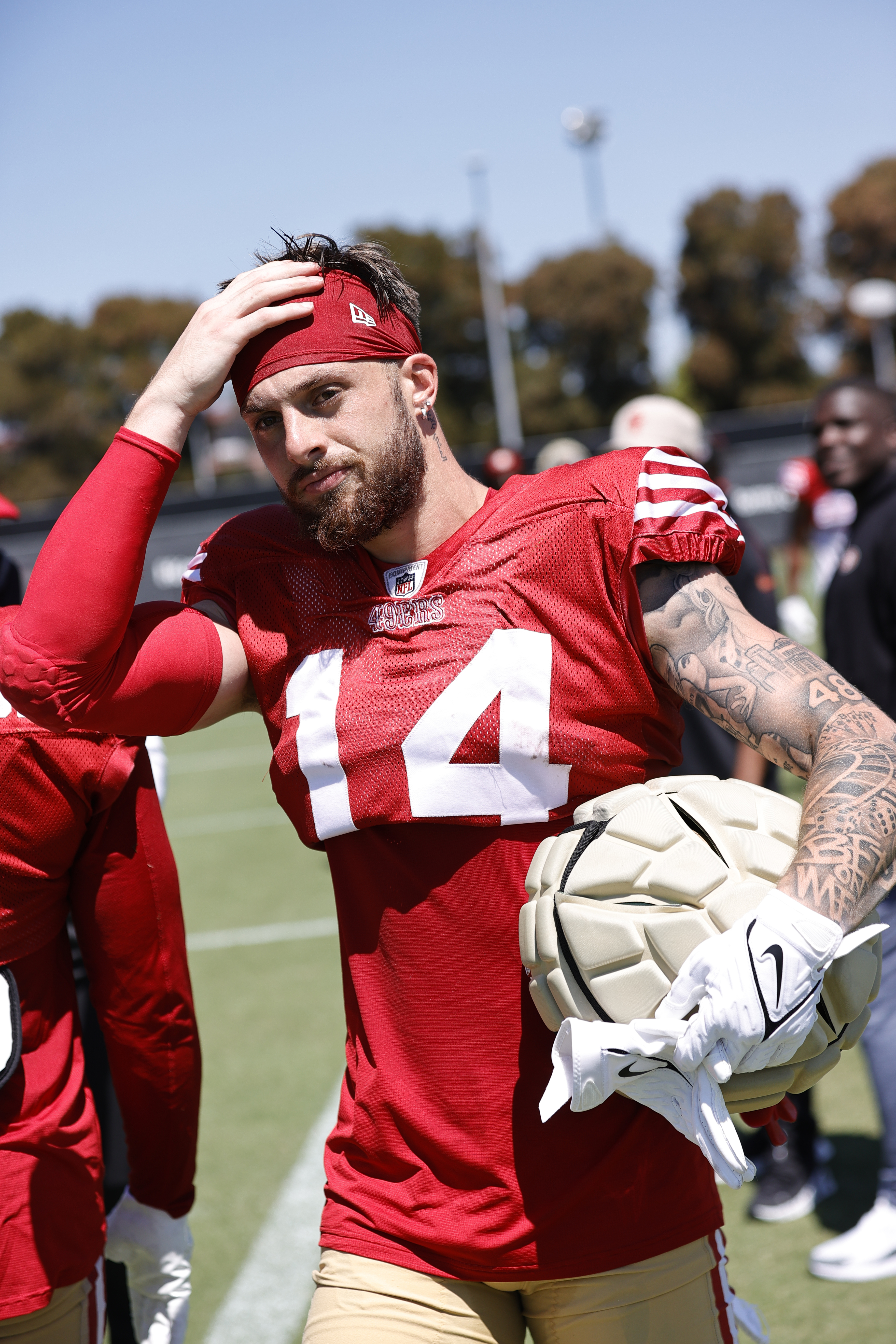 A football player in a red jersey with the number 14 holds a helmet, wipes sweat from his head, and has tattoos on his left arm, standing on a field with people in the background.