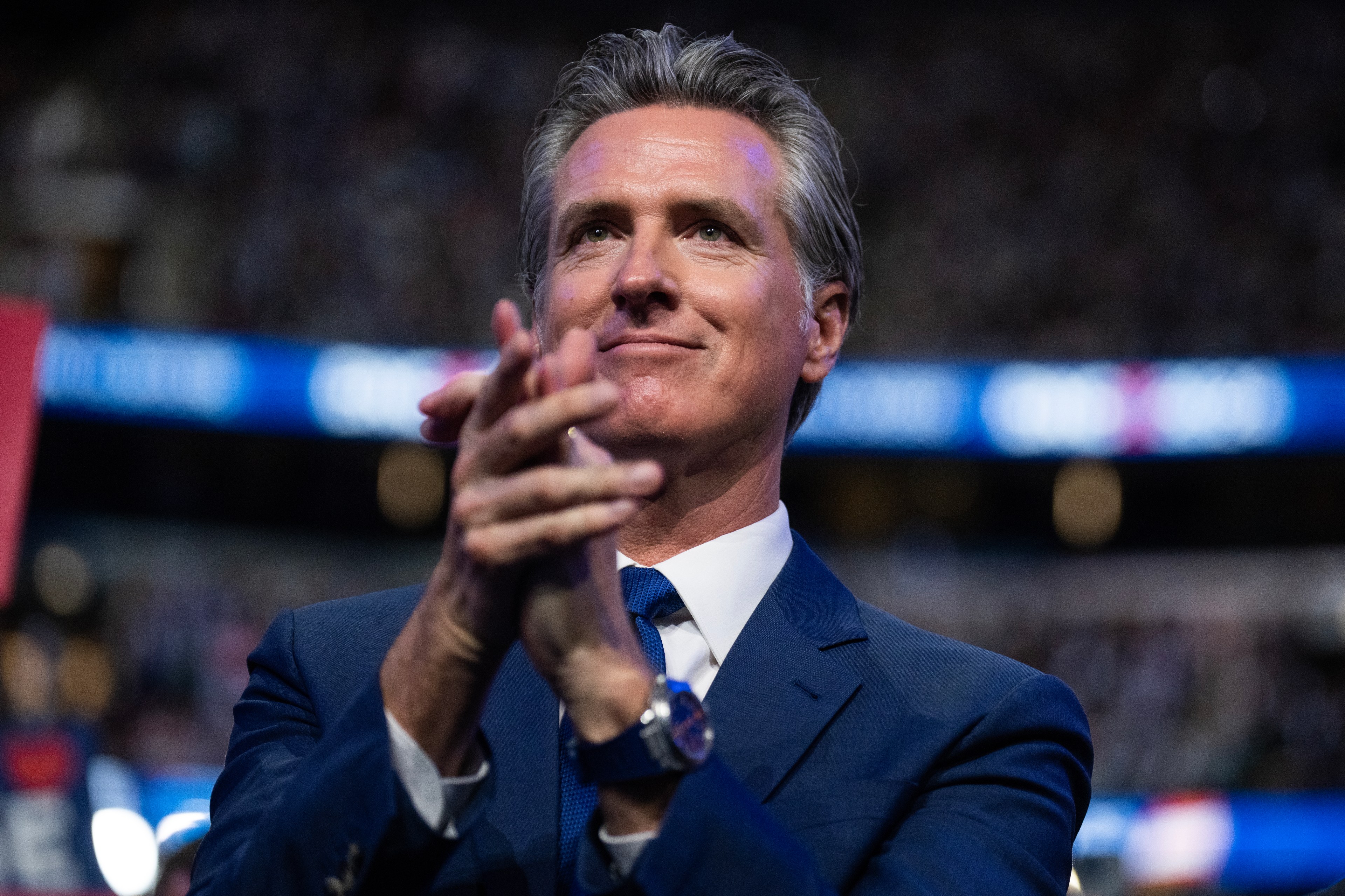 A man in a blue suit claps his hands, smiling slightly. The background is blurred, possibly an indoor event with soft lighting and a blue banner.