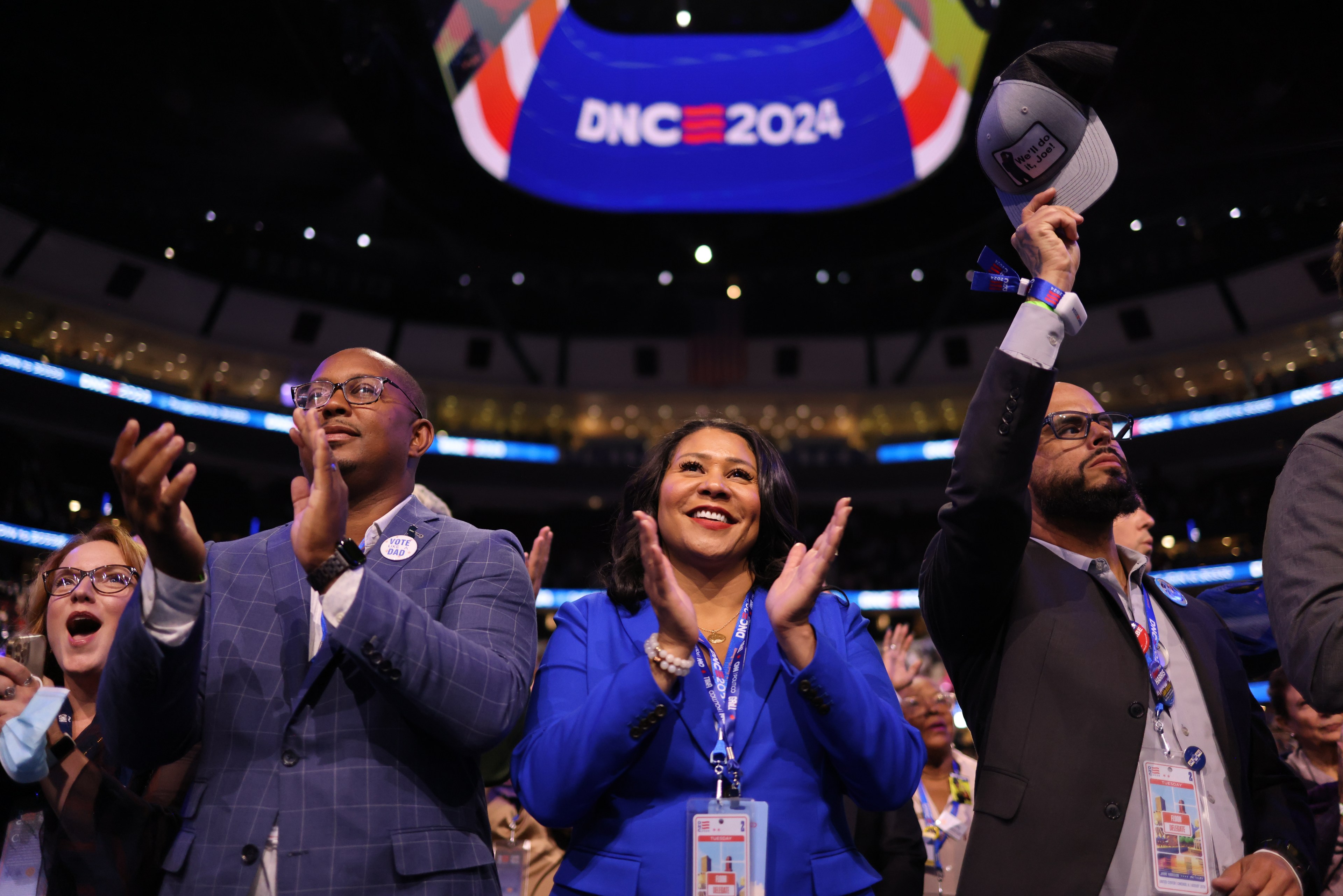 People are clapping and cheering in a crowded convention center. A large &quot;DNC 2024&quot; sign is displayed overhead, creating an energetic atmosphere.