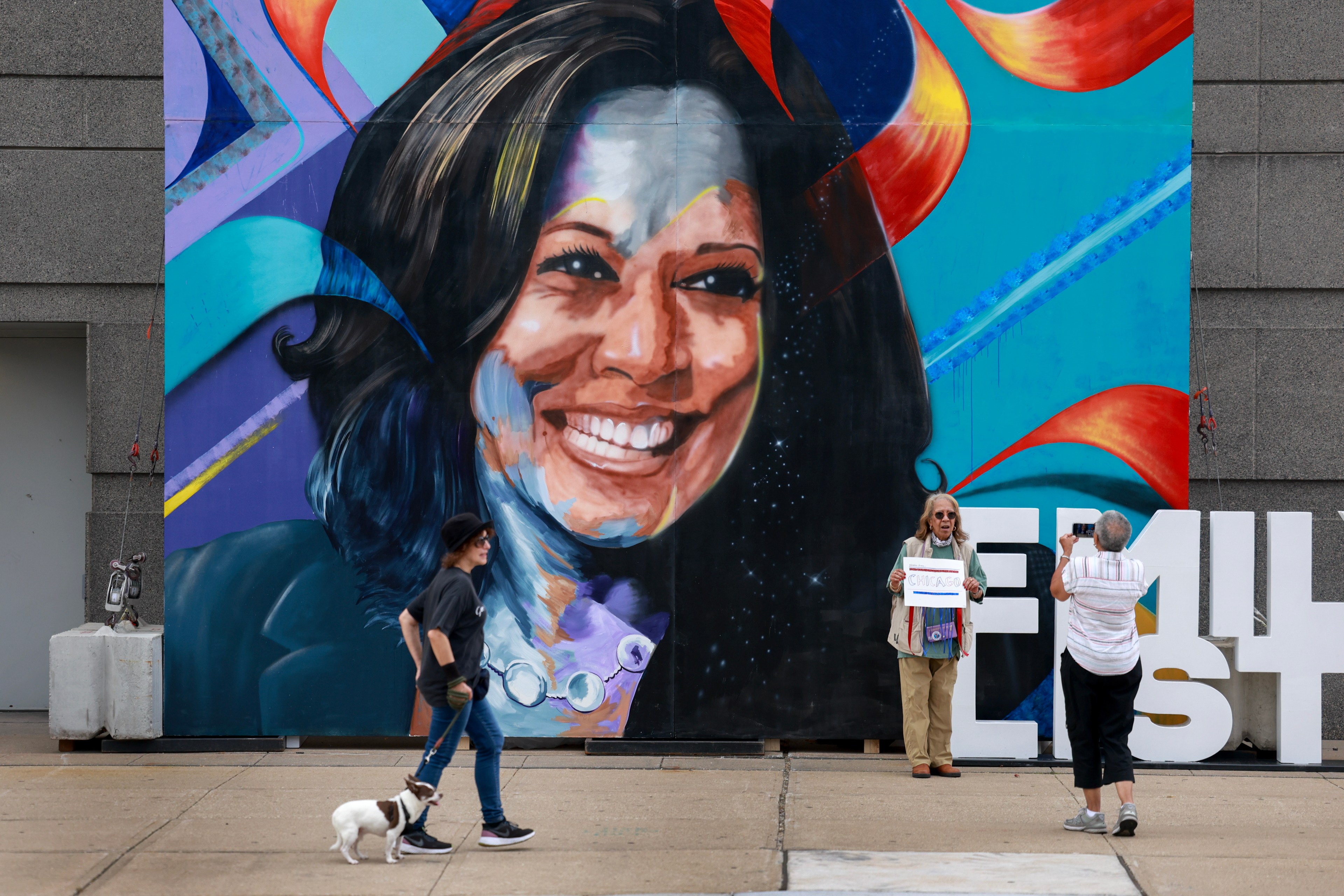 The image shows a large, colorful mural of a smiling woman's face on a wall. In the foreground, a person walks a small dog, and two people take a photo near large white letters.