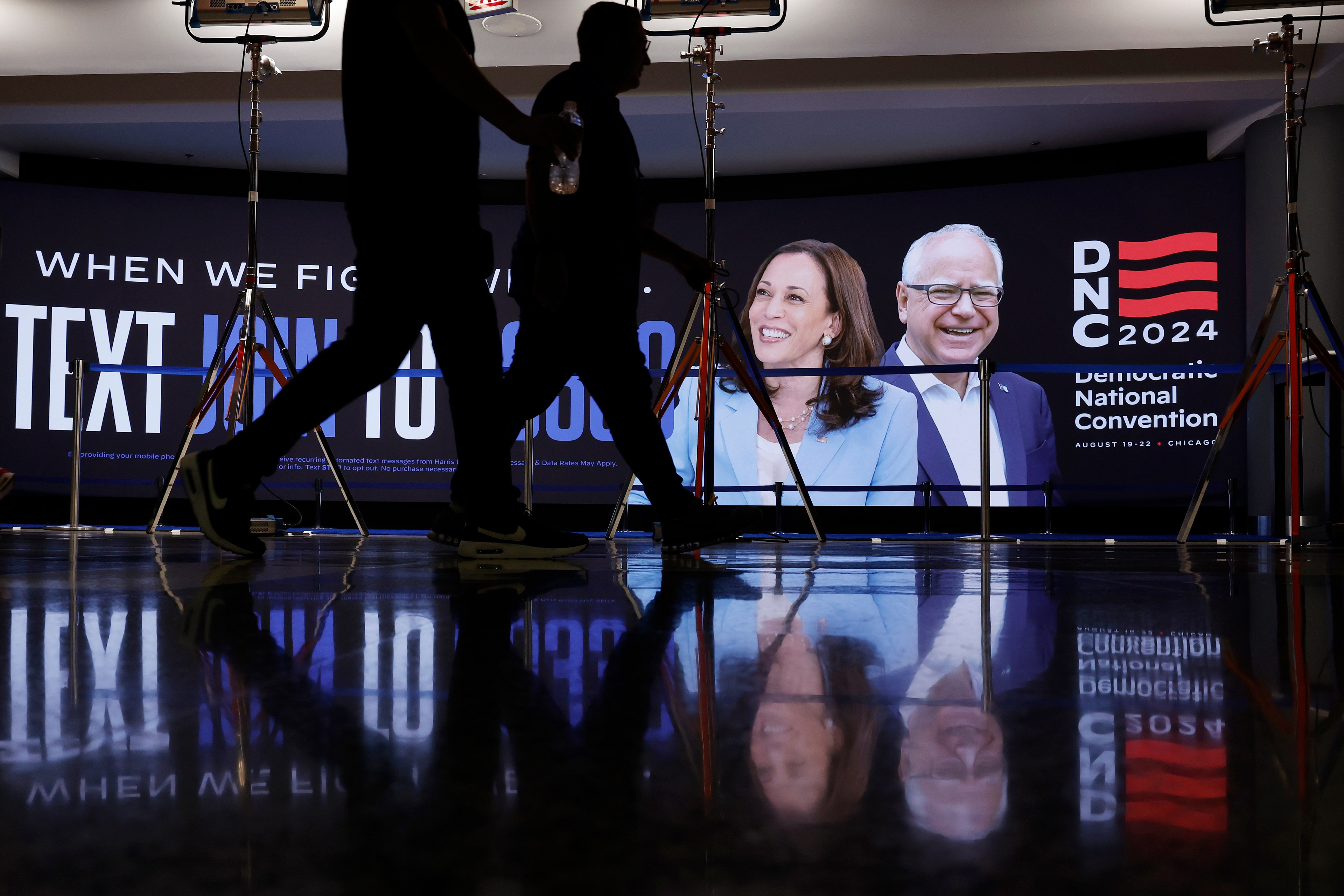 Two people walk past a large screen showing a man and a woman, with &quot;DNC 2024&quot; and event details in the background. Equipment and their reflections are visible.