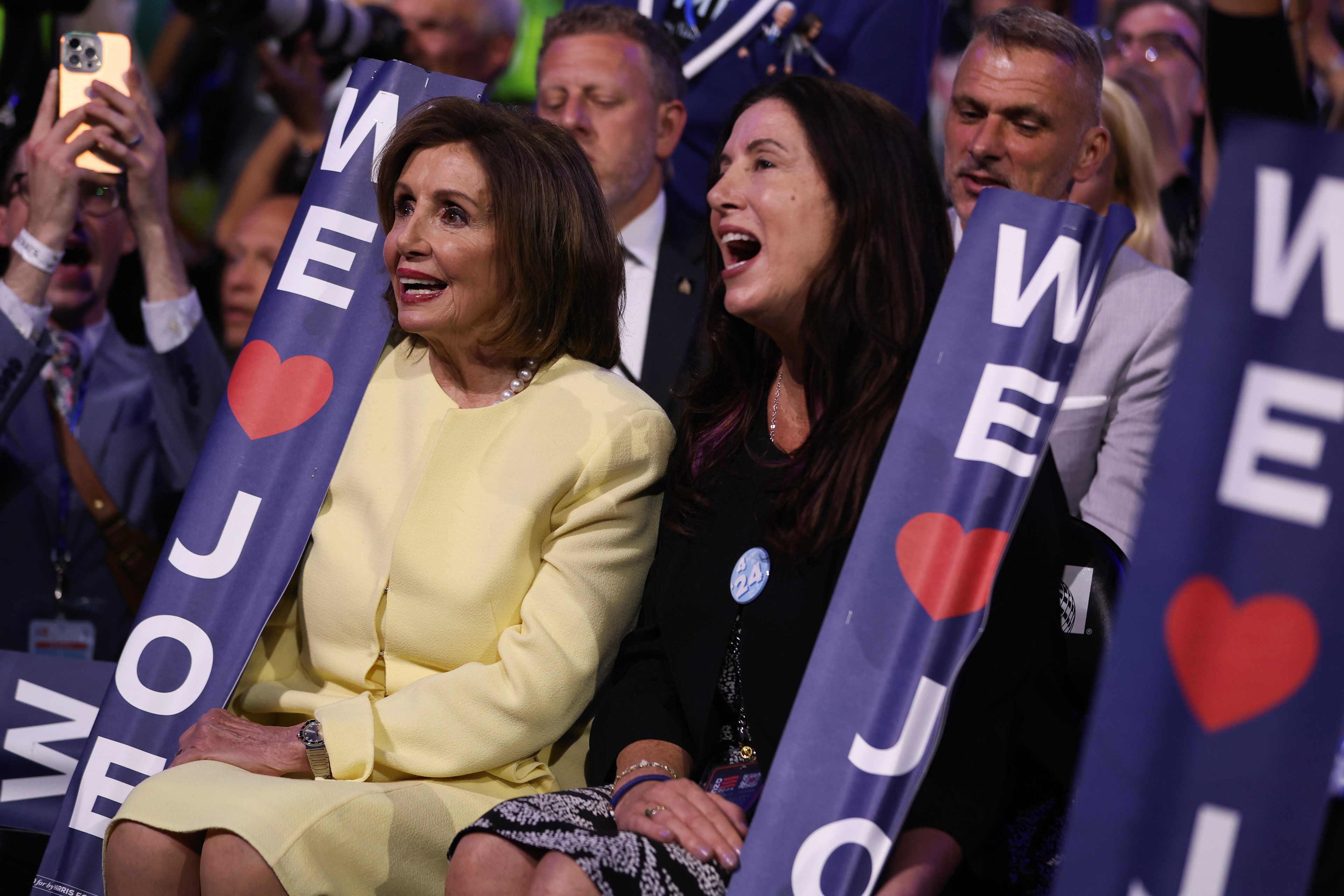 Two women sit smiling among a crowd, holding signs that read &quot;WE ♥ JOE&quot; at a lively event.