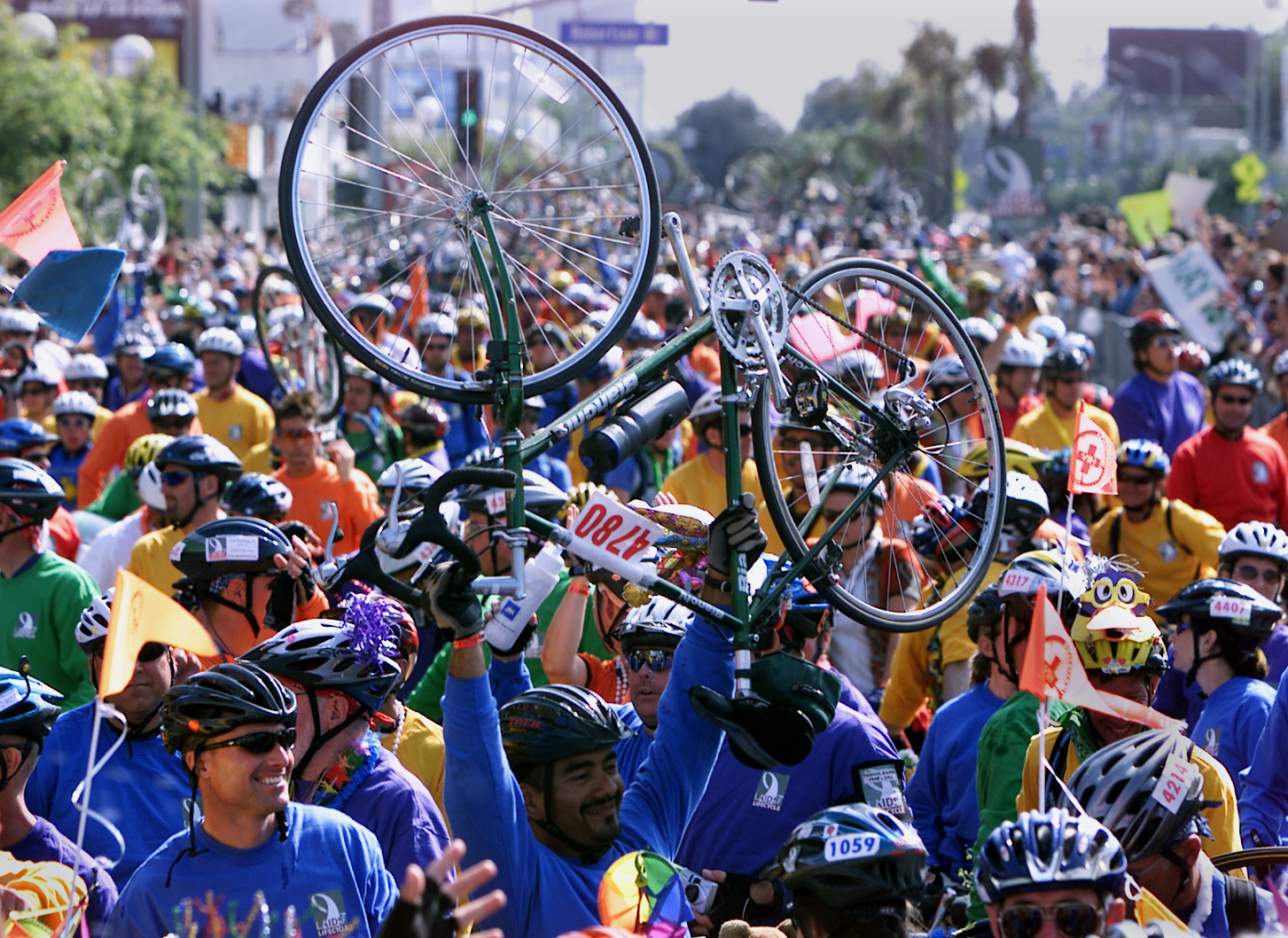 A large, colorful crowd of cyclists in helmets and vibrant outfits are gathered. One person holds up a bicycle above their head, creating a lively and celebratory atmosphere.