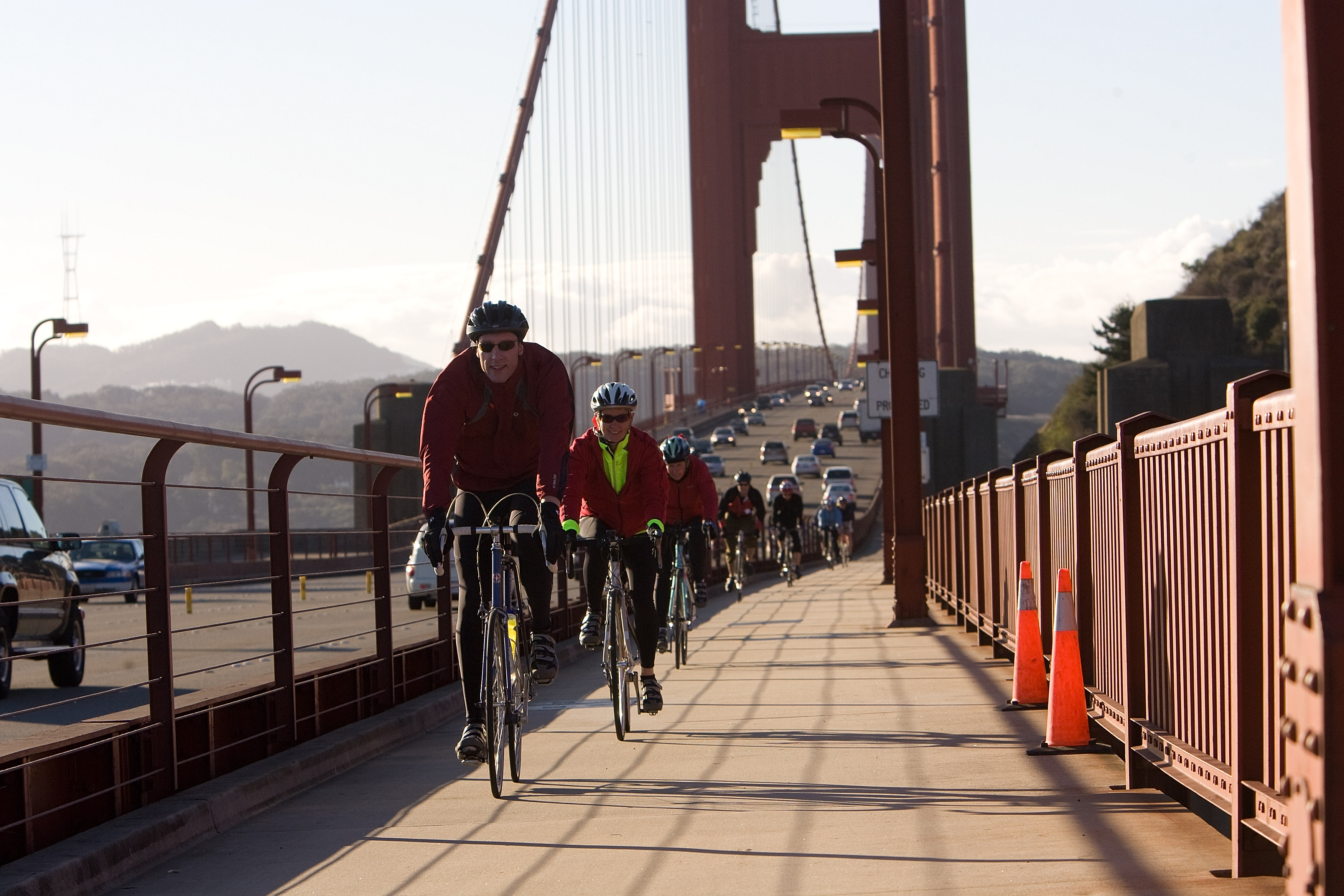 Several cyclists, all wearing helmets and jackets, ride on a pathway alongside a busy bridge with cars. Traffic cones are set on the right railing. Mountains are visible.