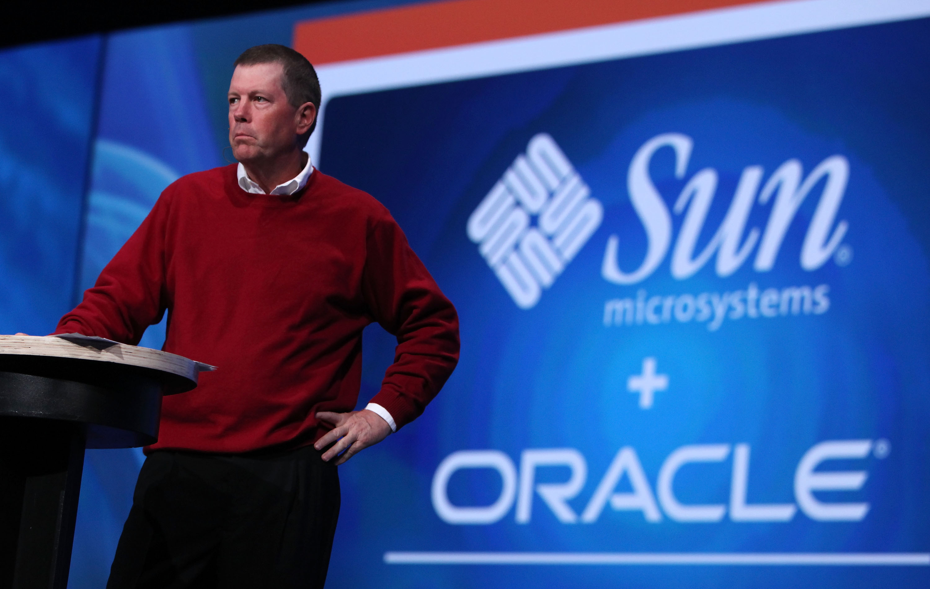 A person in a red sweater stands by a podium, with a large screen behind displaying &quot;Sun Microsystems + Oracle&quot; logos.