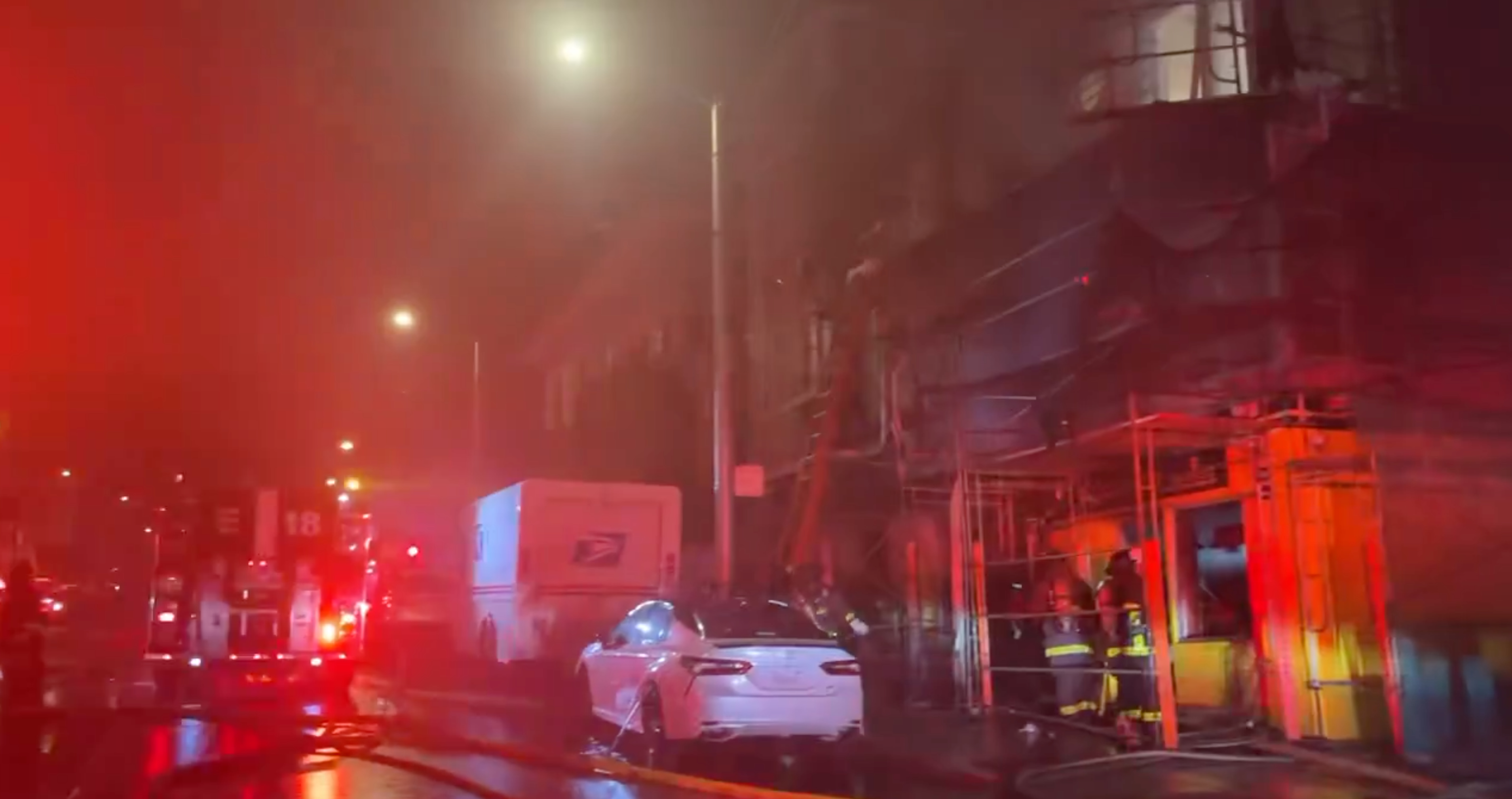 The image shows a nighttime scene with fire trucks, a postal truck, and a white car amid foggy, smoky conditions. Firefighters are actively fighting a building fire.