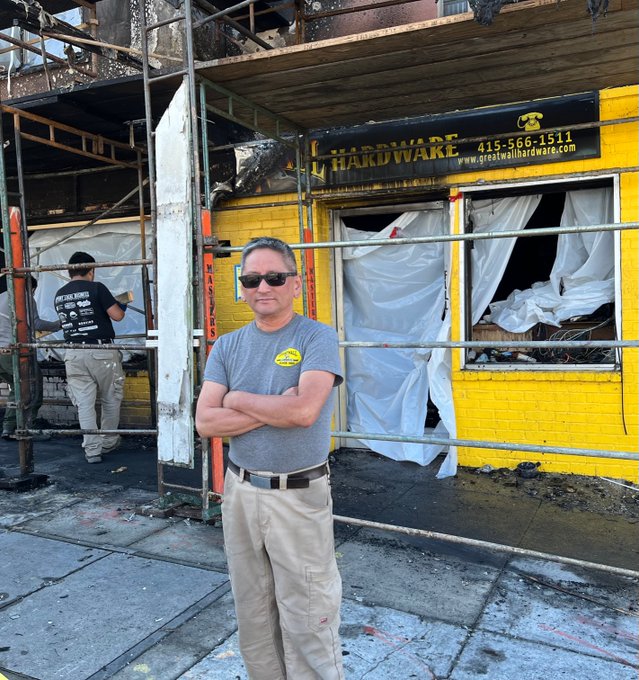 A man in sunglasses stands with arms crossed in front of a burnt yellow hardware store with plastic covering its windows. Another man is working on the damaged store.