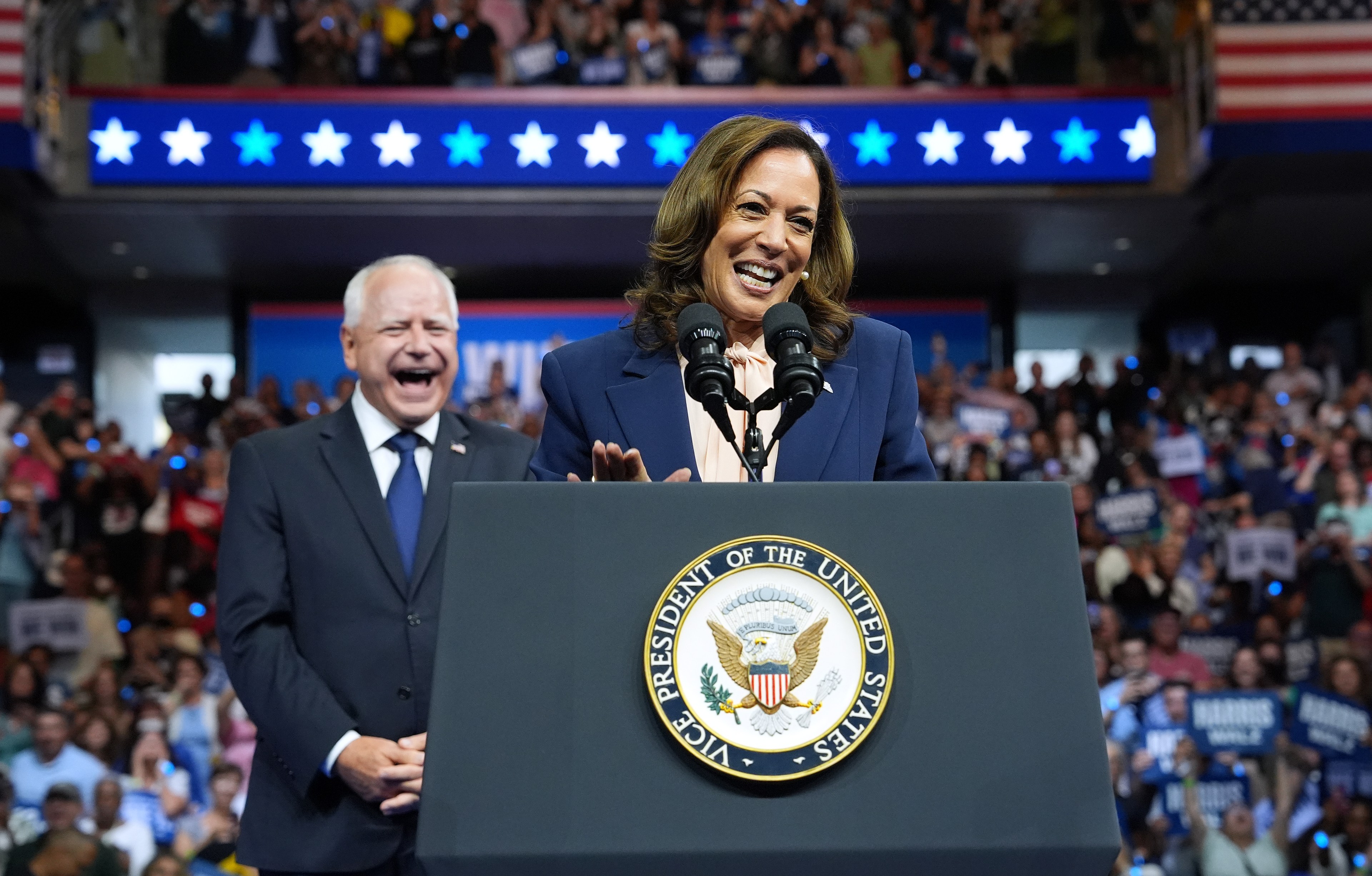 A woman in a suit is speaking at a podium with the Vice President's seal, while a man behind her laughs. A crowd with signs is cheering in the background.