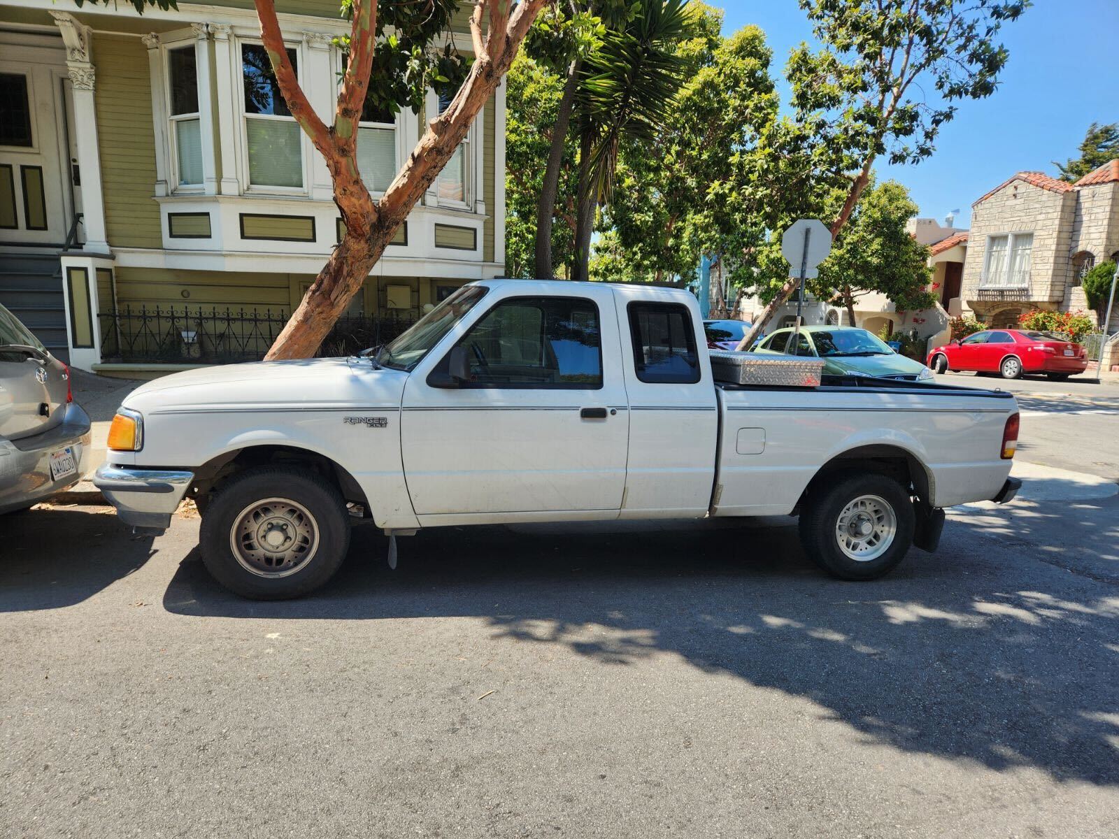 A white pickup truck is parked on the street beside a residential building with bay windows and trees around. Other cars and houses are visible in the background.