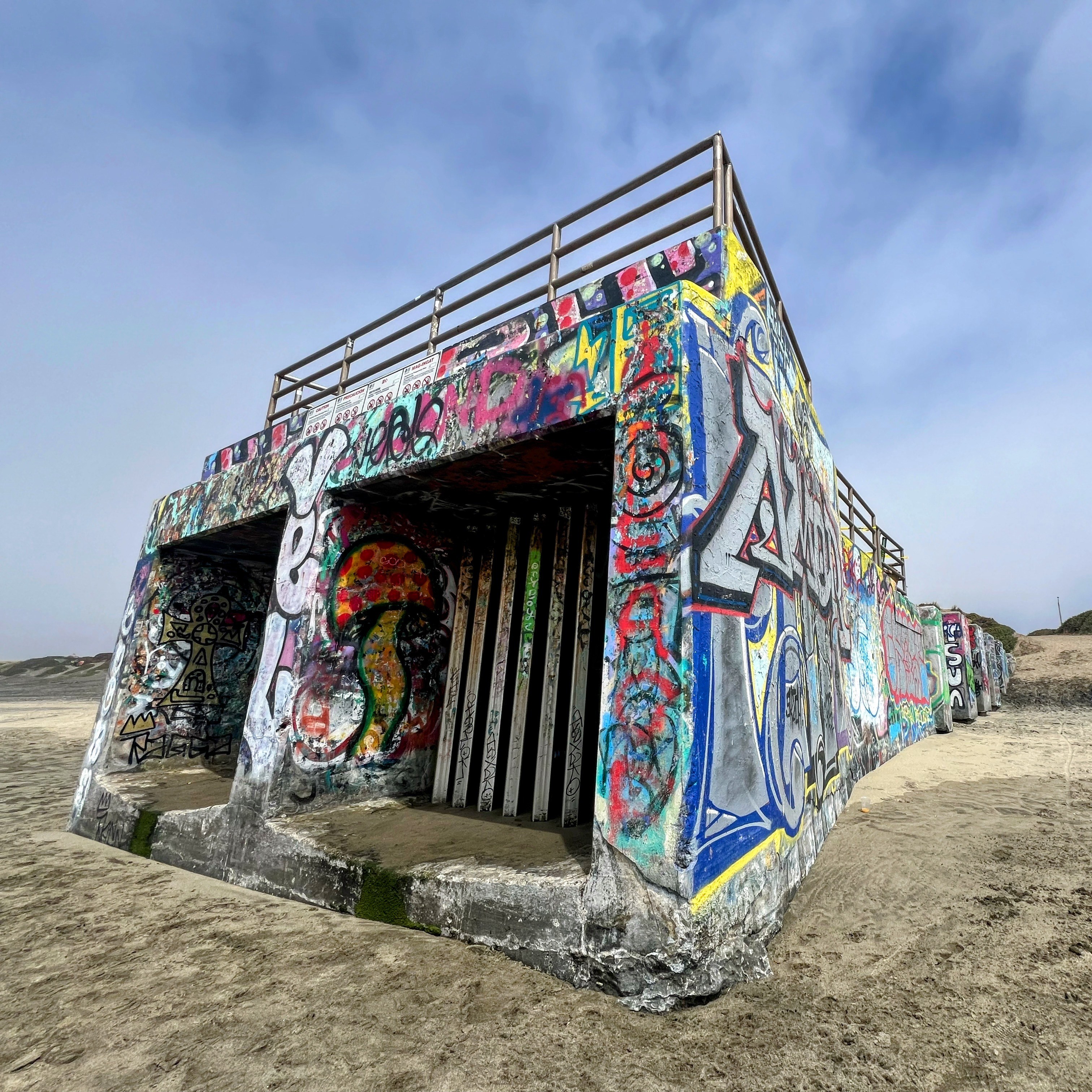 A concrete structure covered in colorful graffiti stands on a sandy beach under a clear, blue sky. The building has two large openings and a fence on the roof.