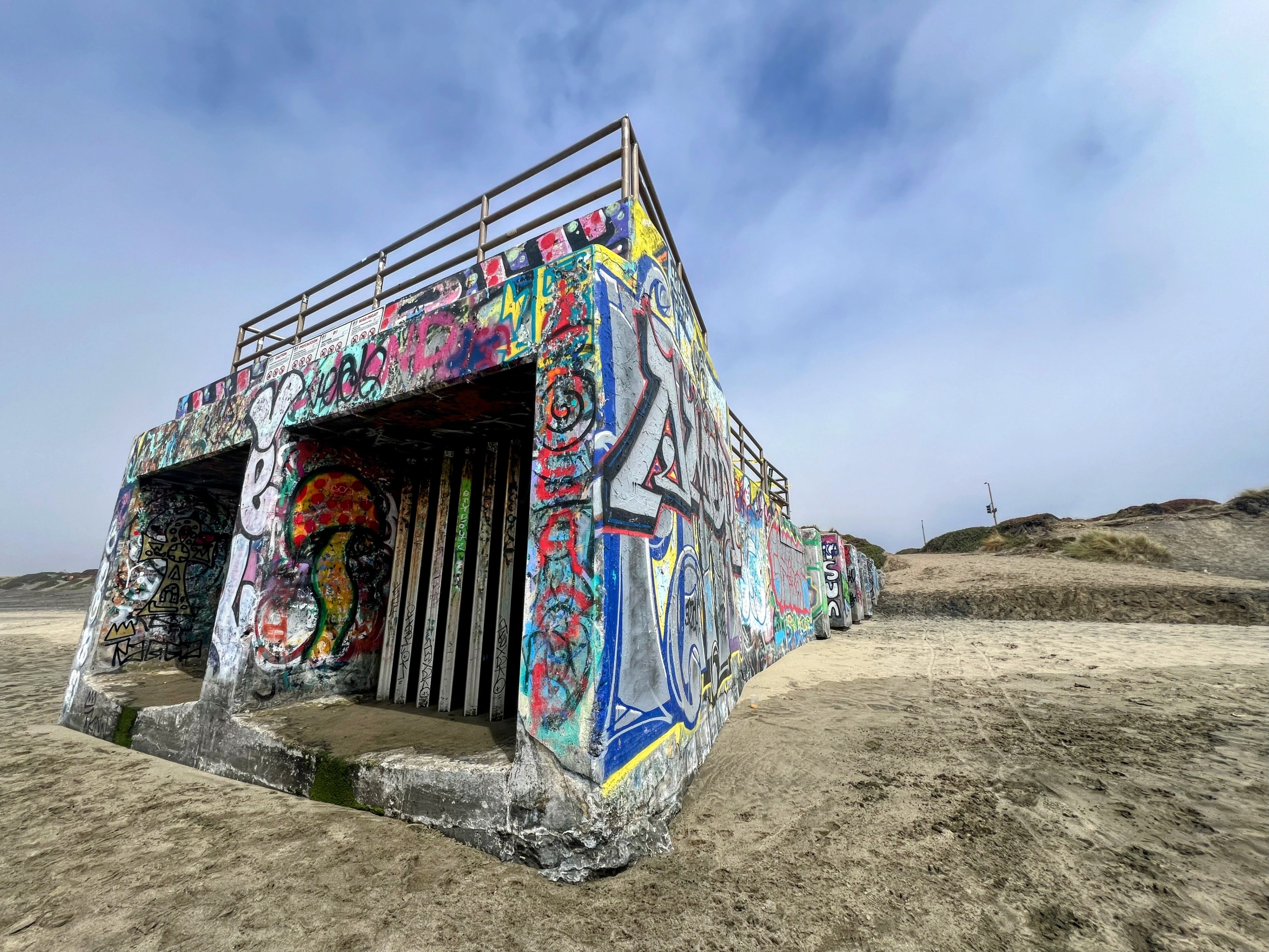 A concrete structure covered in colorful graffiti stands on a sandy beach under a clear, blue sky. The building has two large openings and a fence on the roof.