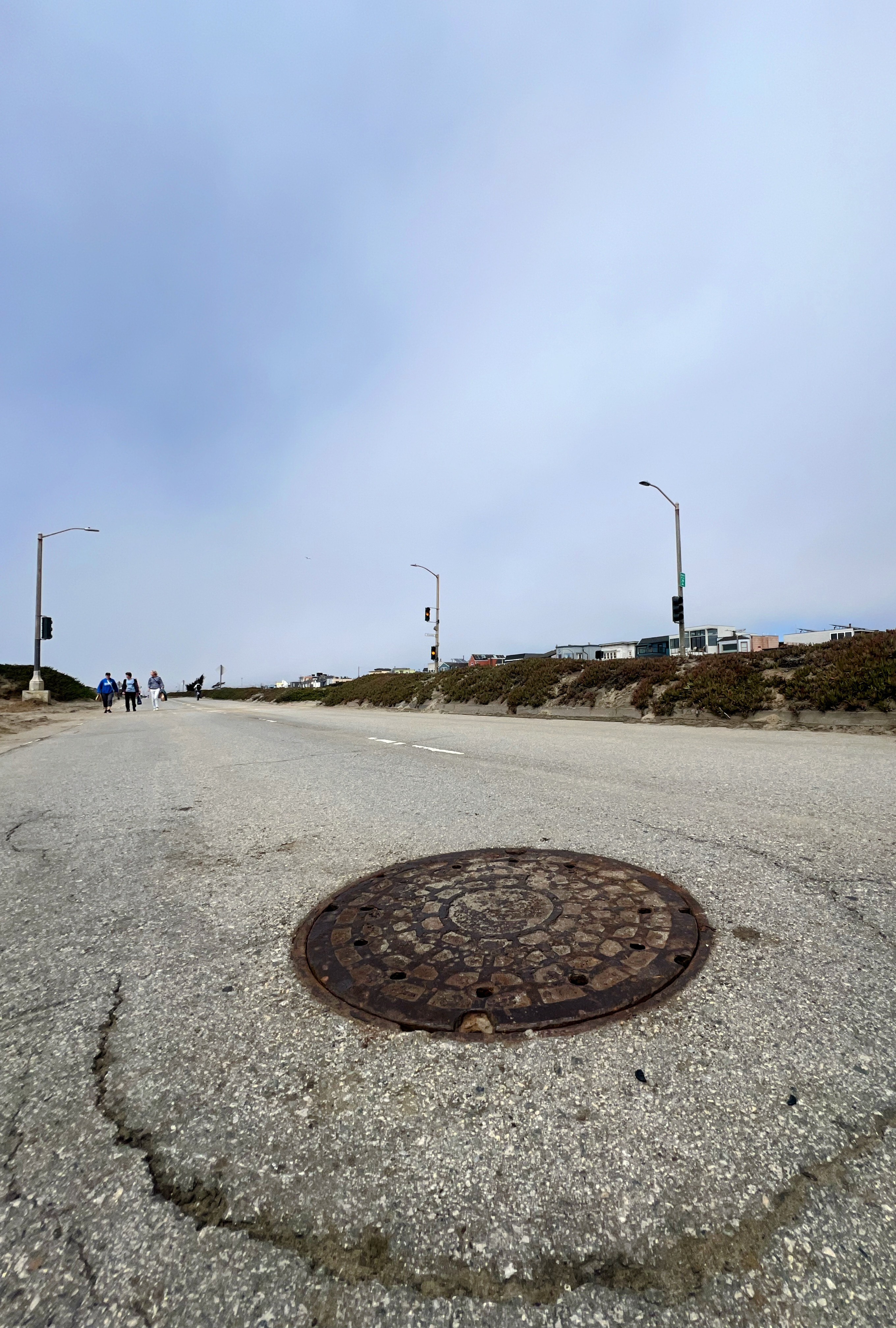 The image shows a manhole cover on a cracked, asphalt road with street lights and people walking in the distance under a cloudy sky.