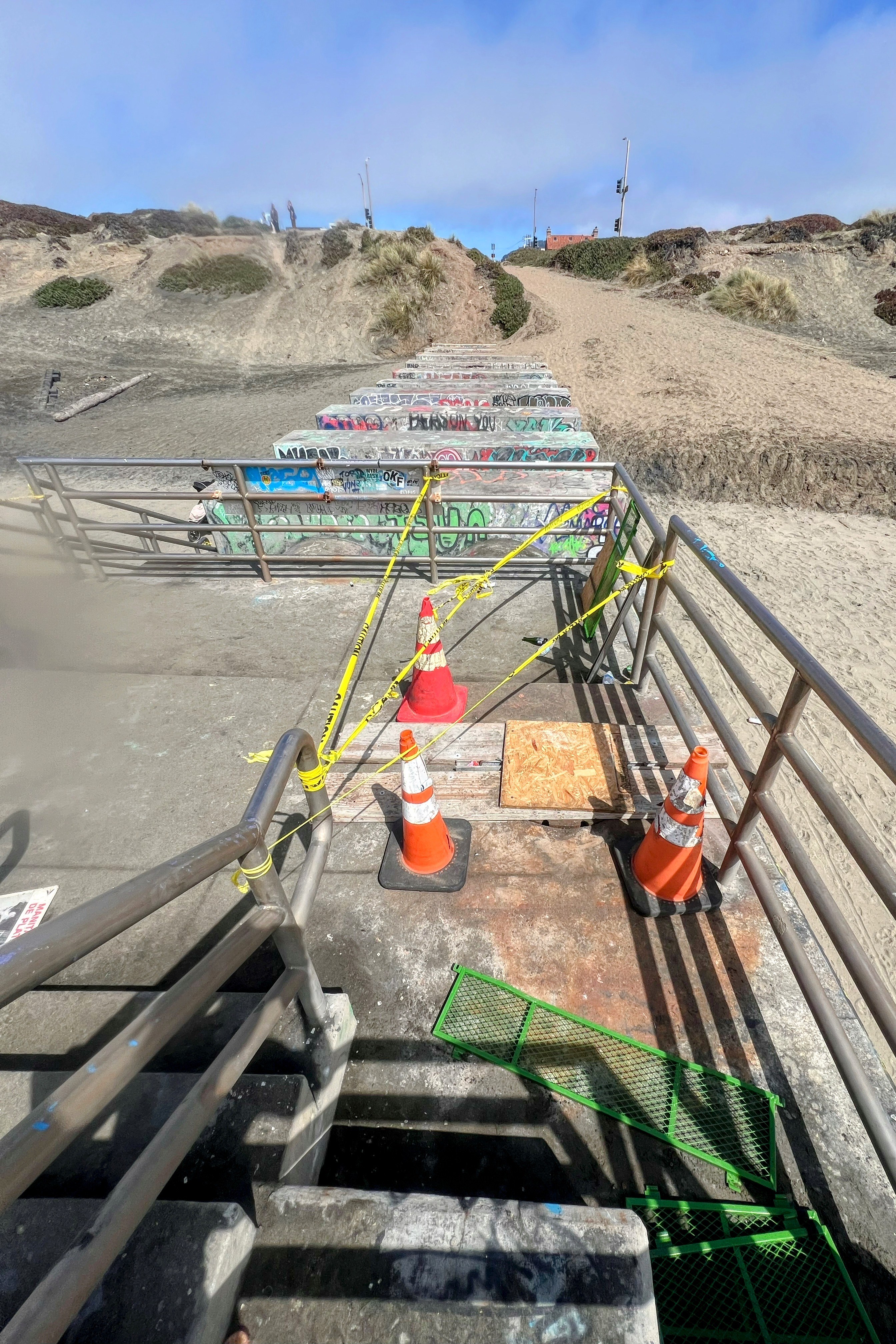 A sandy path leads down to a staircase covered in graffiti, with sections blocked off by orange cones and caution tape. Some metal grating is displaced.