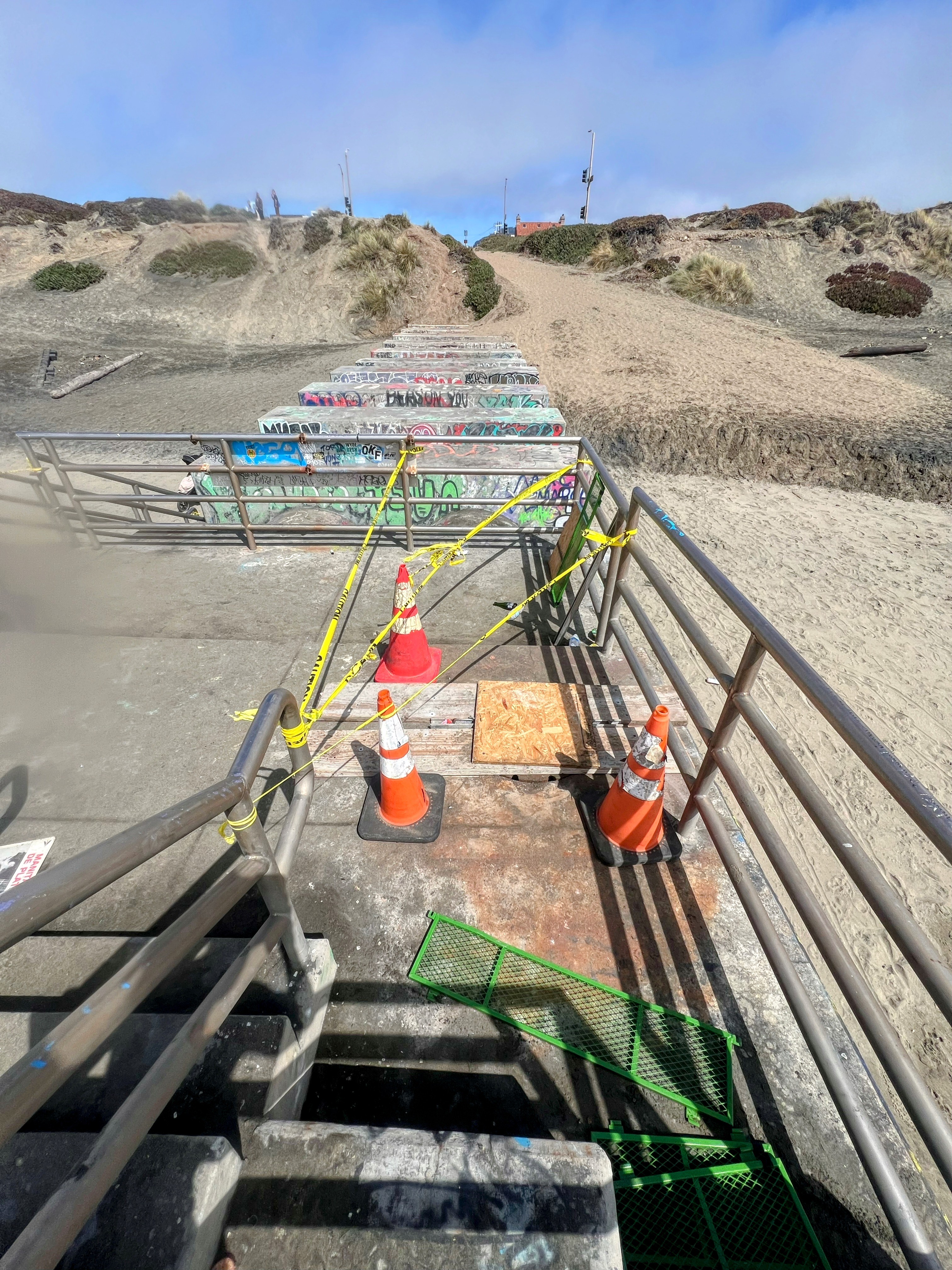 A sandy path leads down to a staircase covered in graffiti, with sections blocked off by orange cones and caution tape. Some metal grating is displaced.