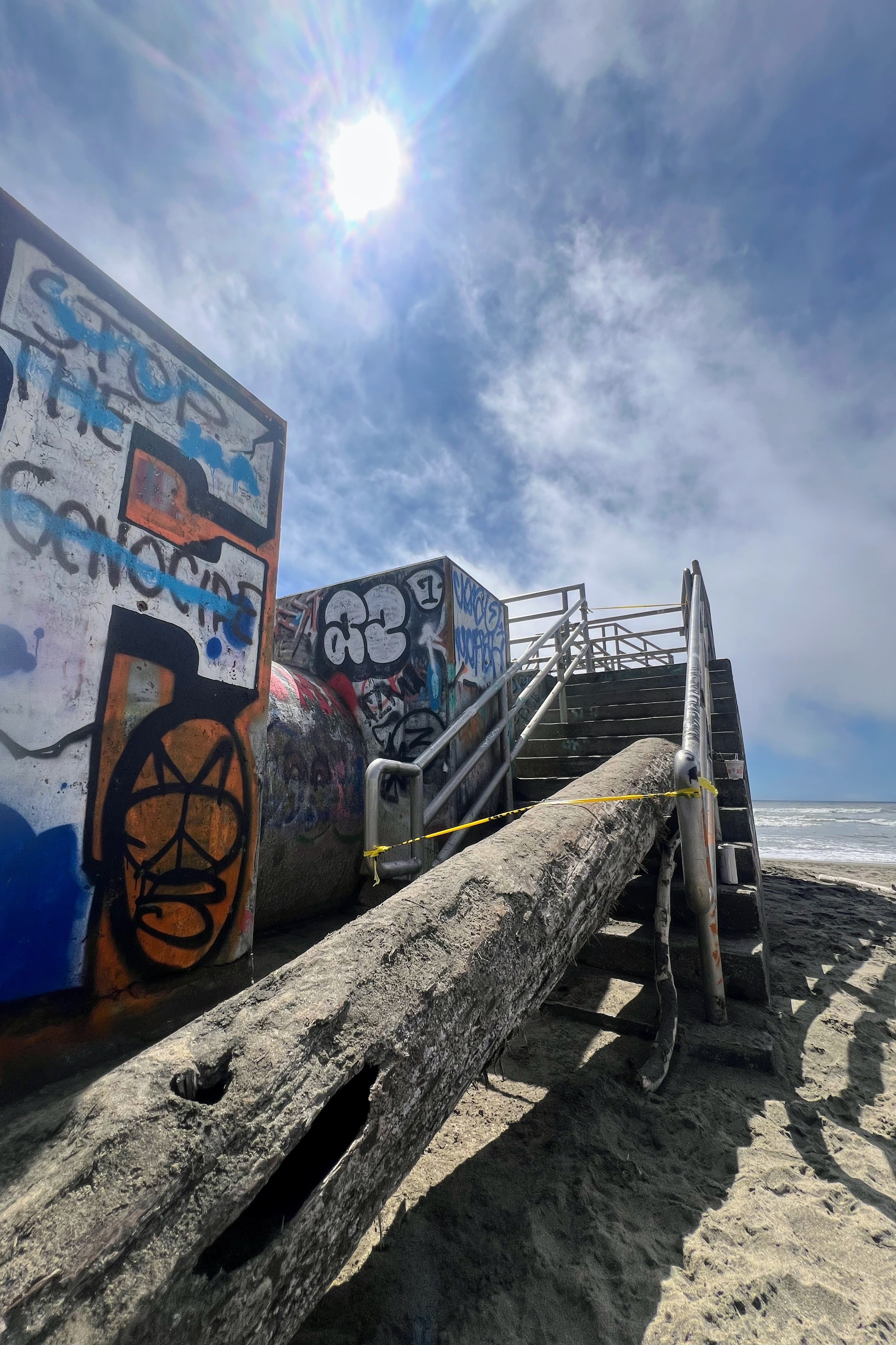 The image shows a sandy beach with graffiti-covered structures and a staircase blocked by a large log, under a bright sun and a partly cloudy sky.