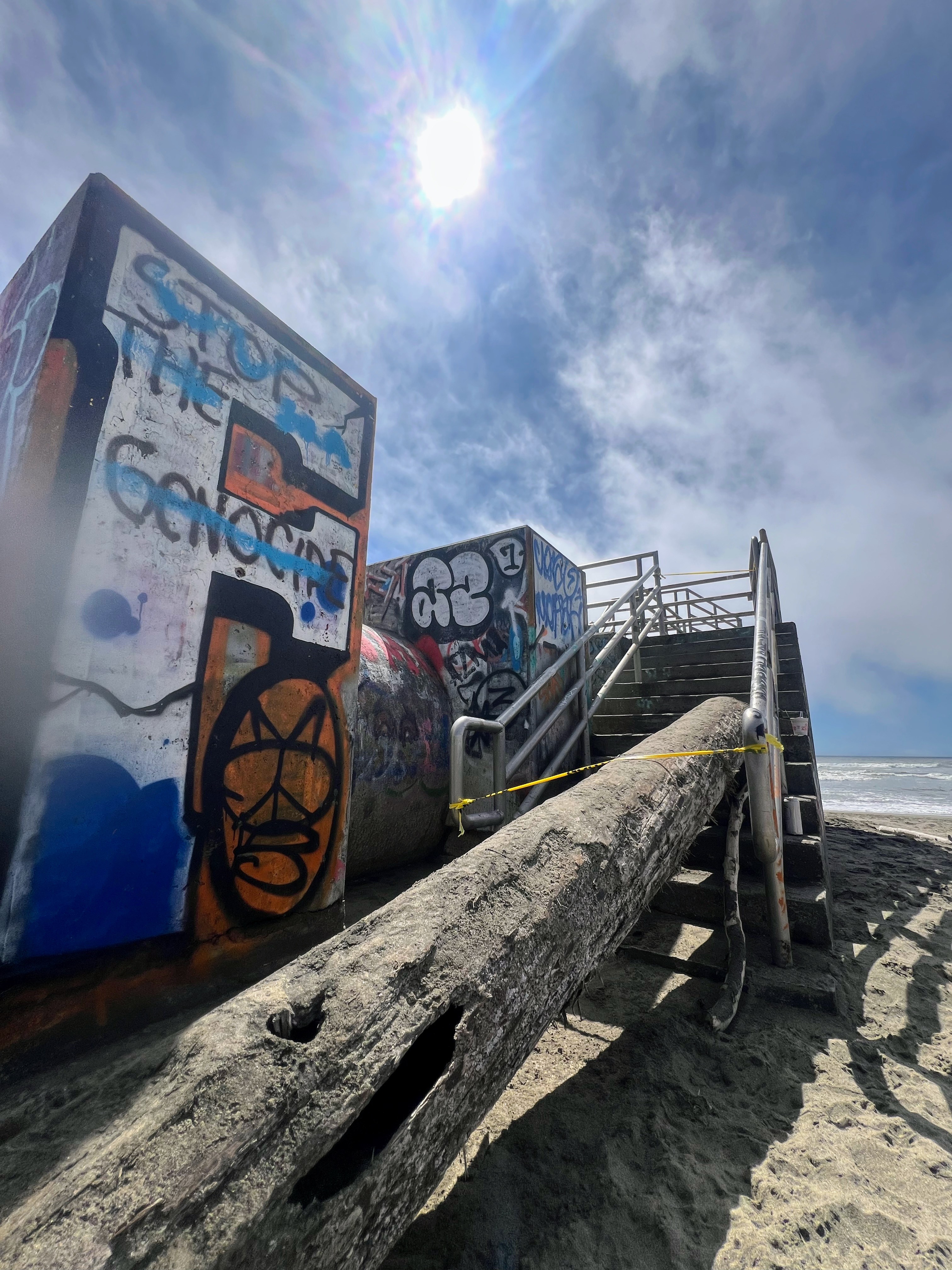 The image shows a sandy beach with graffiti-covered structures and a staircase blocked by a large log, under a bright sun and a partly cloudy sky.