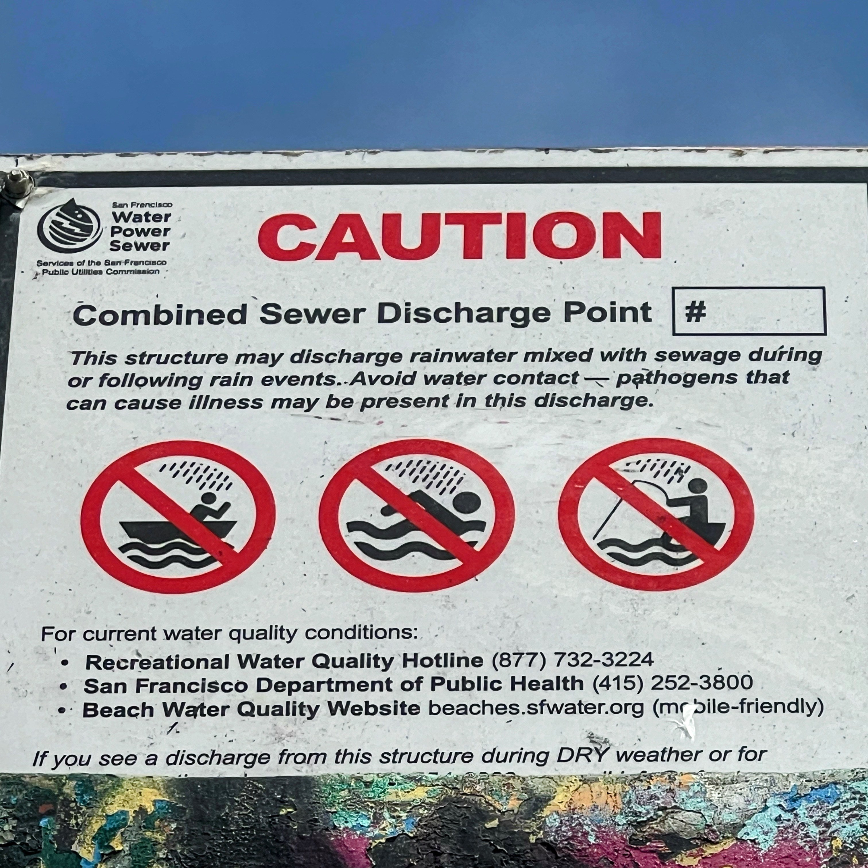 A warning sign indicating a combined sewer discharge point, cautioning against water contact due to possible rainwater and sewage mixing, with contact details for water quality conditions.