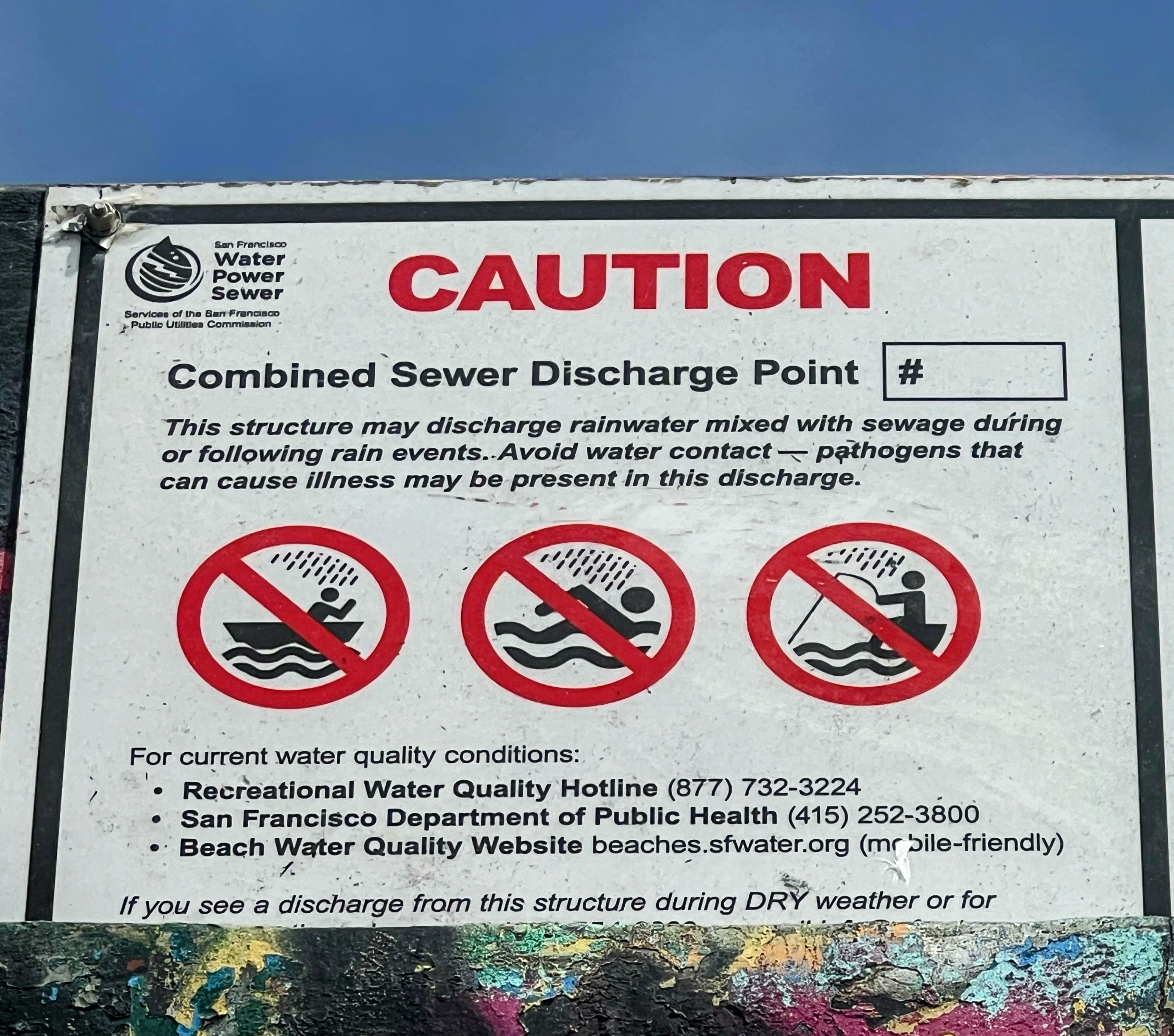 A warning sign indicating a combined sewer discharge point, cautioning against water contact due to possible rainwater and sewage mixing, with contact details for water quality conditions.