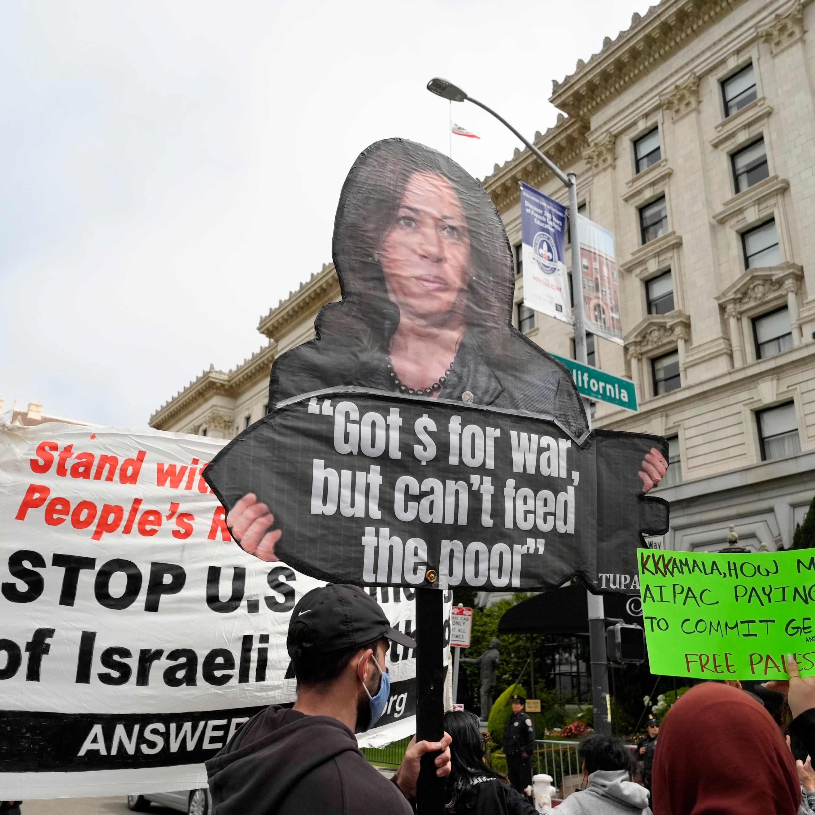 Protesters hold various signs, including one with a picture of a person and the text &quot;Got $ for war, but can't feed the poor&quot; and others showing support for Palestine.