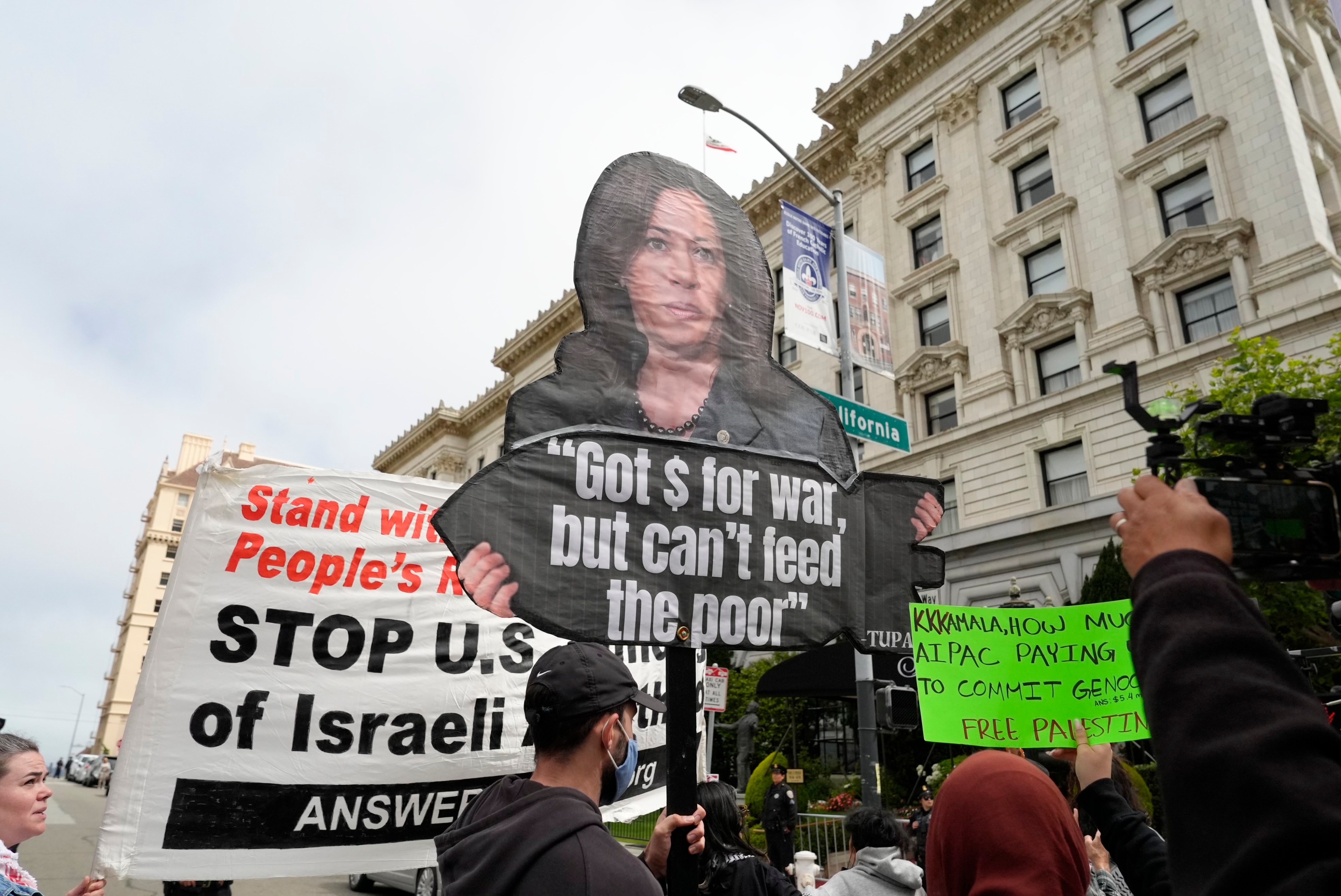 Protesters hold various signs, including one with a picture of a person and the text &quot;Got $ for war, but can't feed the poor&quot; and others showing support for Palestine.