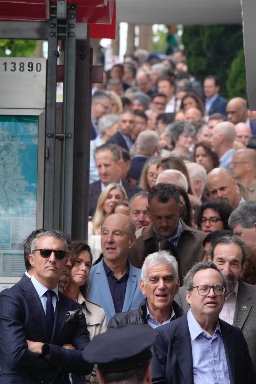 A long line of people stand closely packed on a sidewalk, extending into the distance, with most individuals looking serious or contemplative.