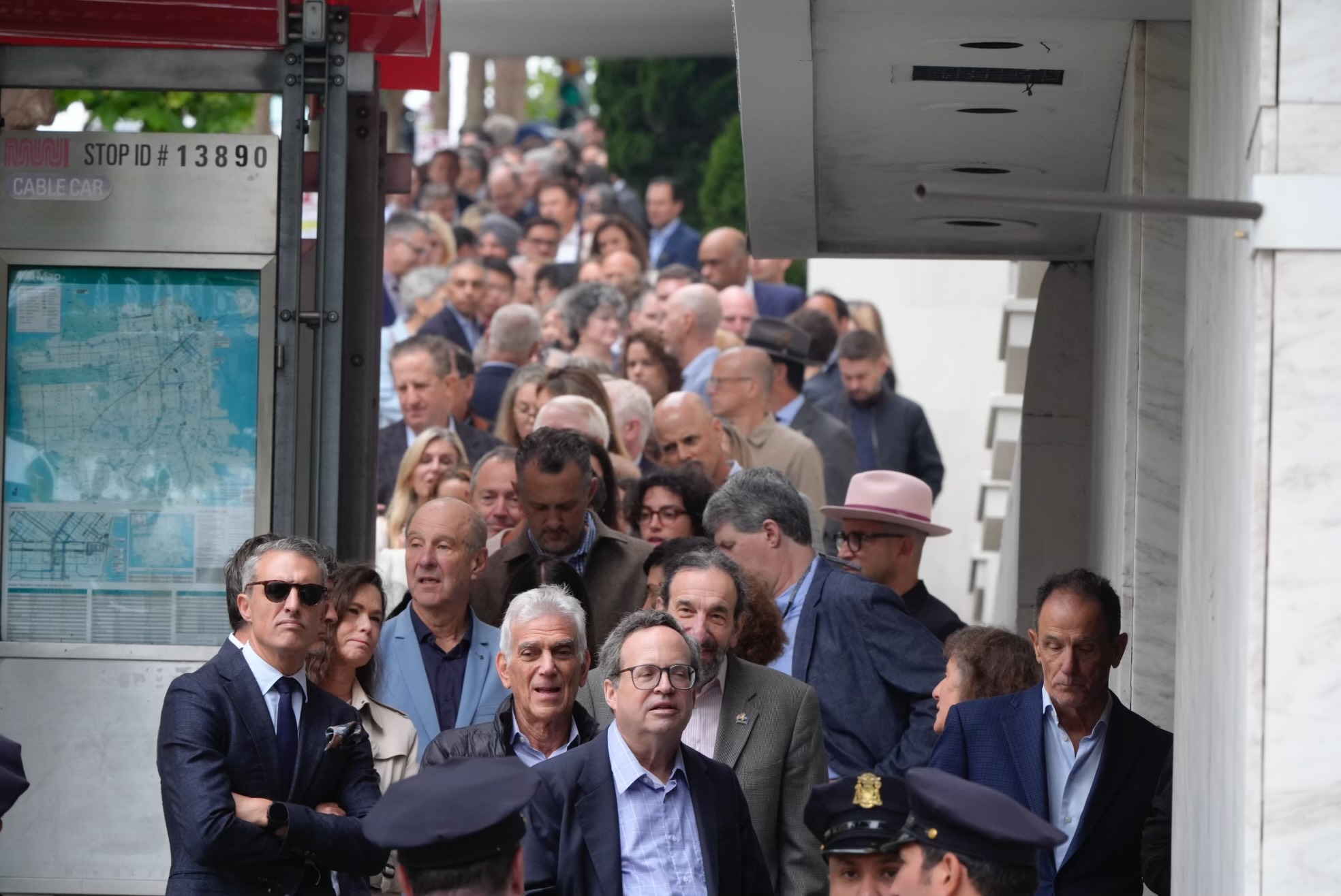 A long line of people stand closely packed on a sidewalk, extending into the distance, with most individuals looking serious or contemplative.