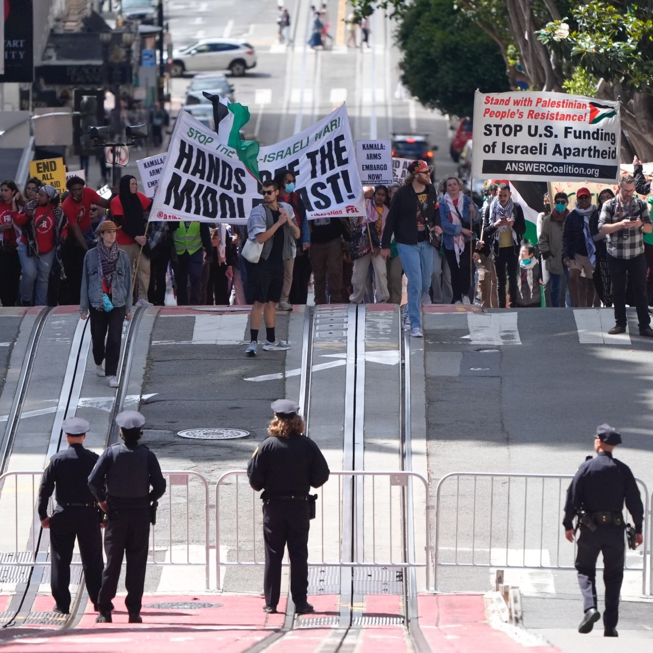 A group of protesters with signs and flags stand behind barricades on a city street, while police officers face them, maintaining a formed line across the road.