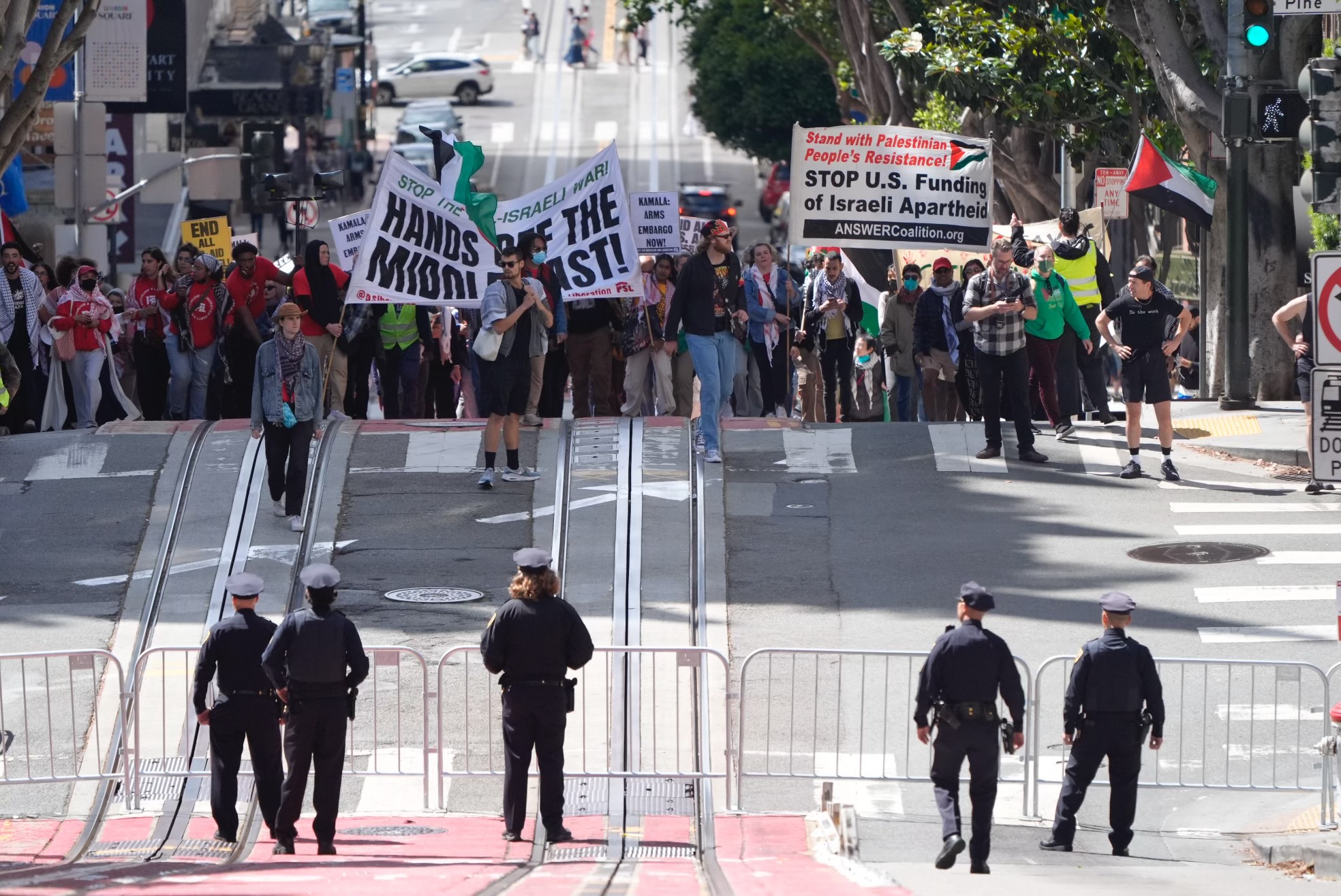 A group of protesters with signs and flags stand behind barricades on a city street, while police officers face them, maintaining a formed line across the road.