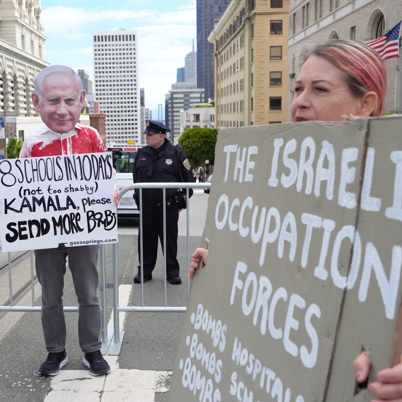 The image shows a protest with people holding signs criticizing Israeli actions. One person wears a mask while others display signs about bombings and occupation.