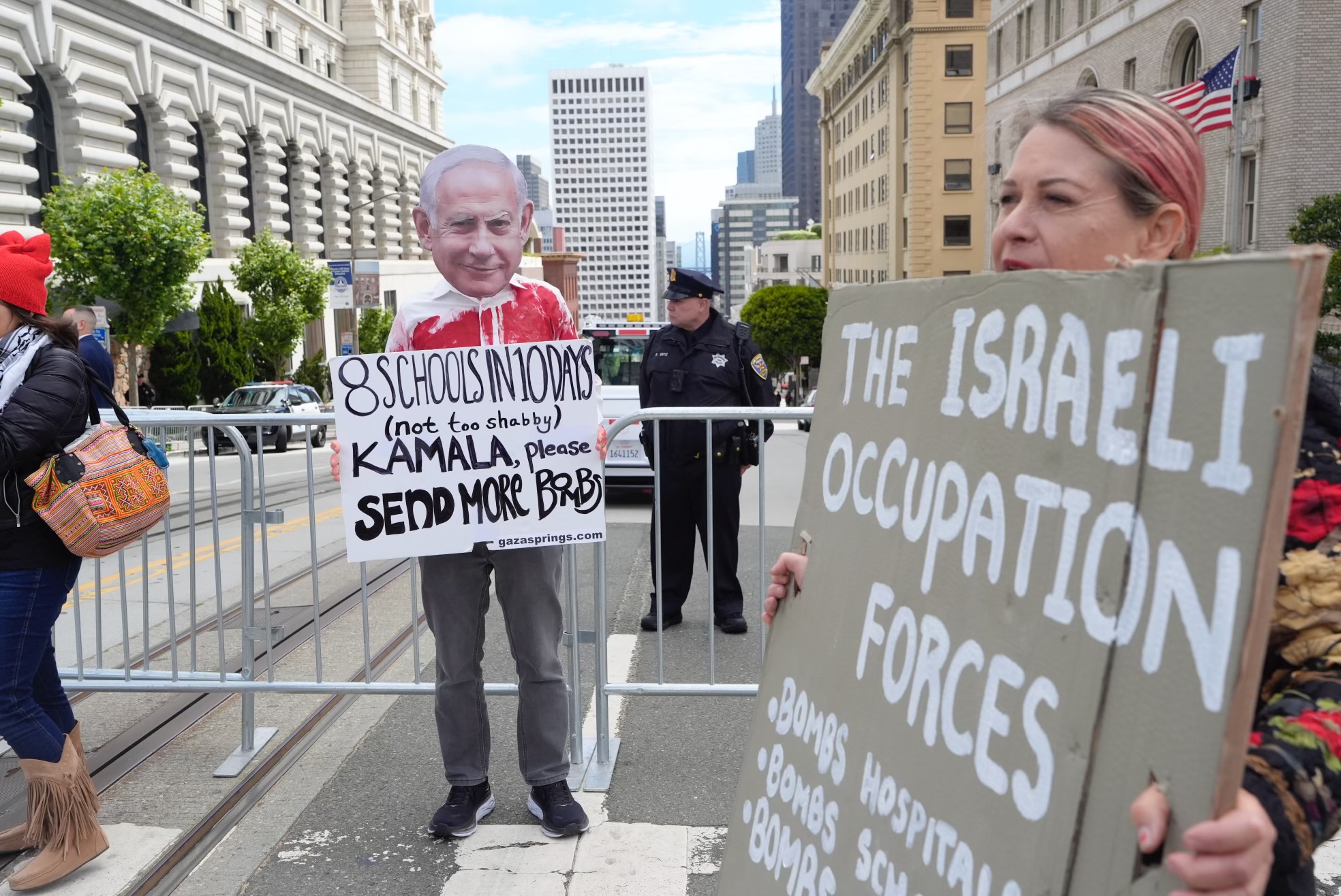 The image shows a protest with people holding signs criticizing Israeli actions. One person wears a mask while others display signs about bombings and occupation.