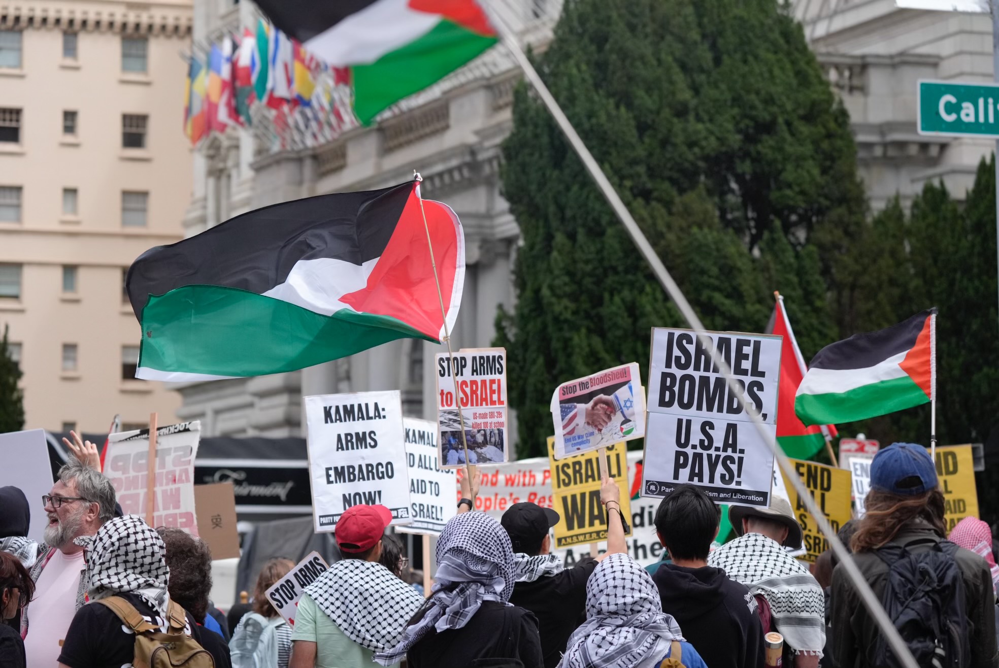 Protesters holding Palestinian flags and signs such as &quot;Stop Arms to Israel&quot; and &quot;Israel Bombs, U.S.A Pays!&quot; gather near a building and intersection named &quot;California&quot;.