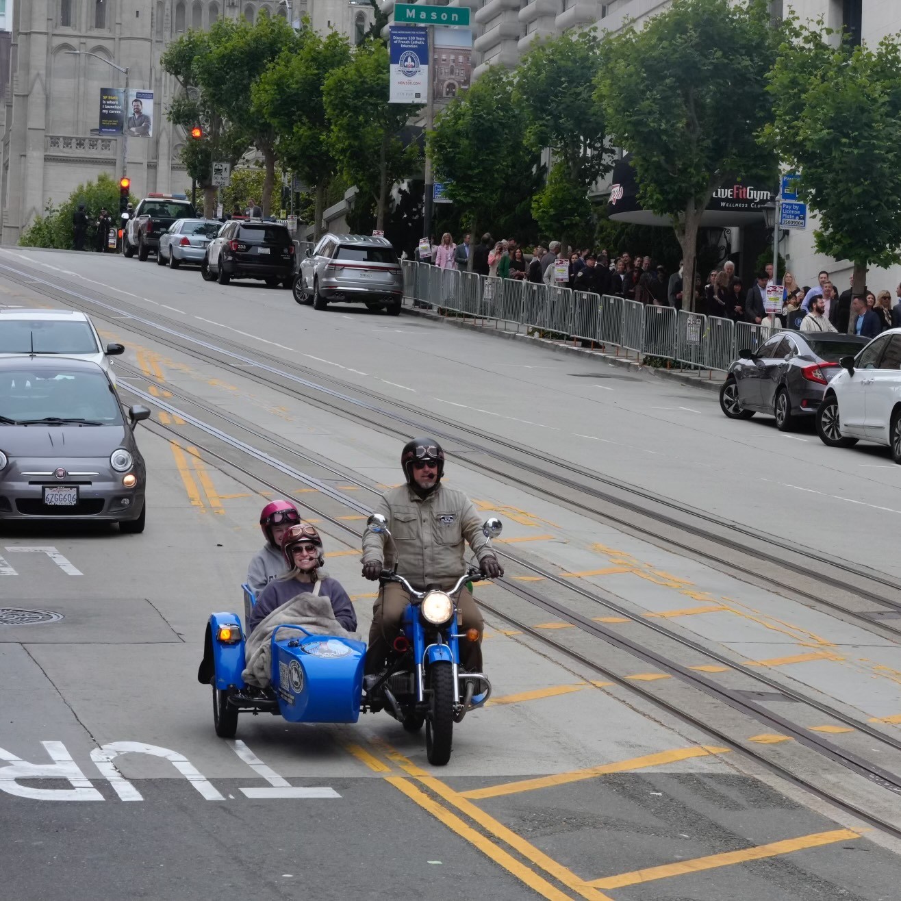 A man rides a blue motorcycle with a sidecar carrying two passengers up a hilly street, while a crowd stands behind barriers on the sidewalk, and cars are parked along the road.