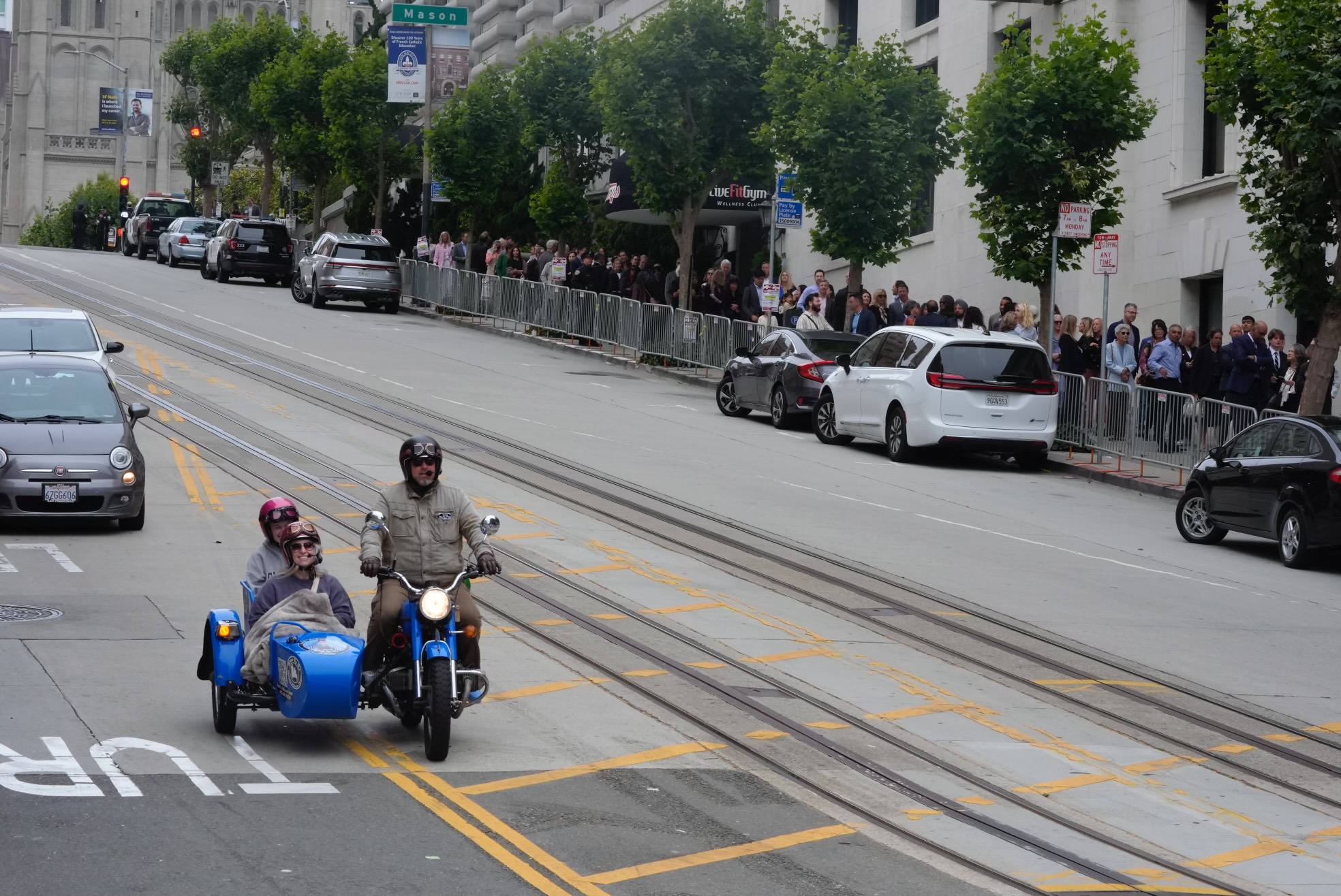 A man rides a blue motorcycle with a sidecar carrying two passengers up a hilly street, while a crowd stands behind barriers on the sidewalk, and cars are parked along the road.