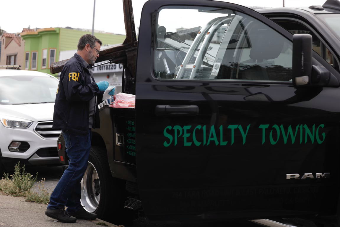 An FBI agent wearing gloves examines items in the open back of a black "Specialty Towing" truck parked on a street next to white and green buildings.