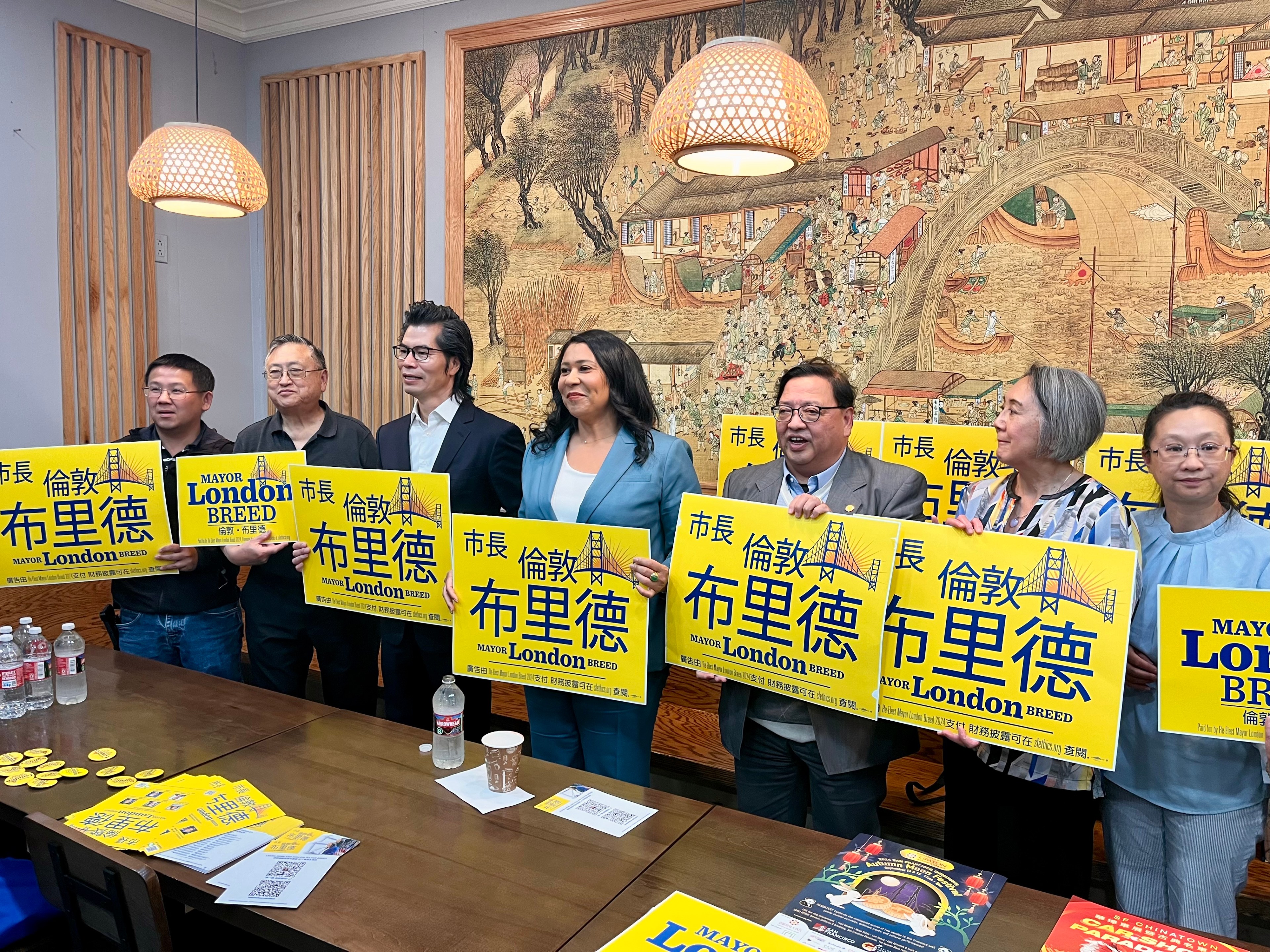 A group of seven people are standing indoors, holding yellow signs that say &quot;Mayor London Breed&quot; in English and Chinese characters. They are smiling and appear supportive.