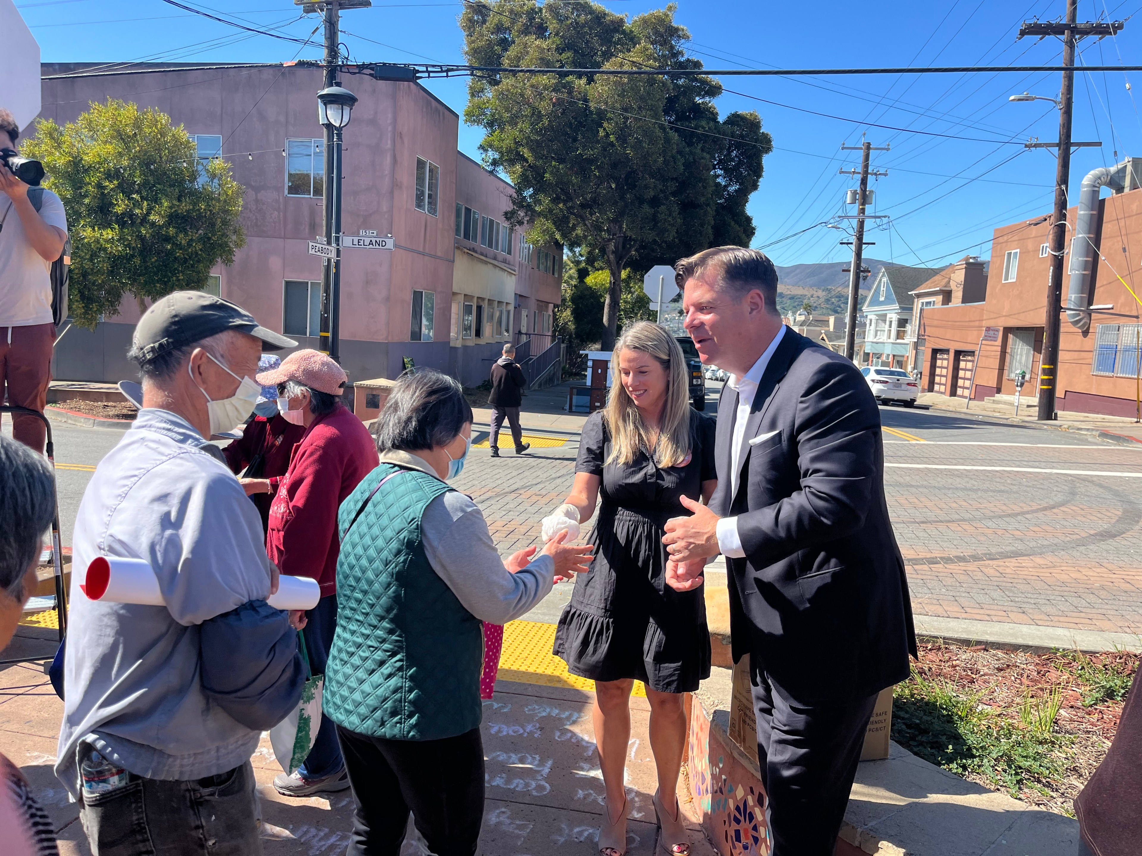 A man in a suit and a woman in a black dress shake hands with two elderly people outdoors; several others stand nearby on a sunny street corner.