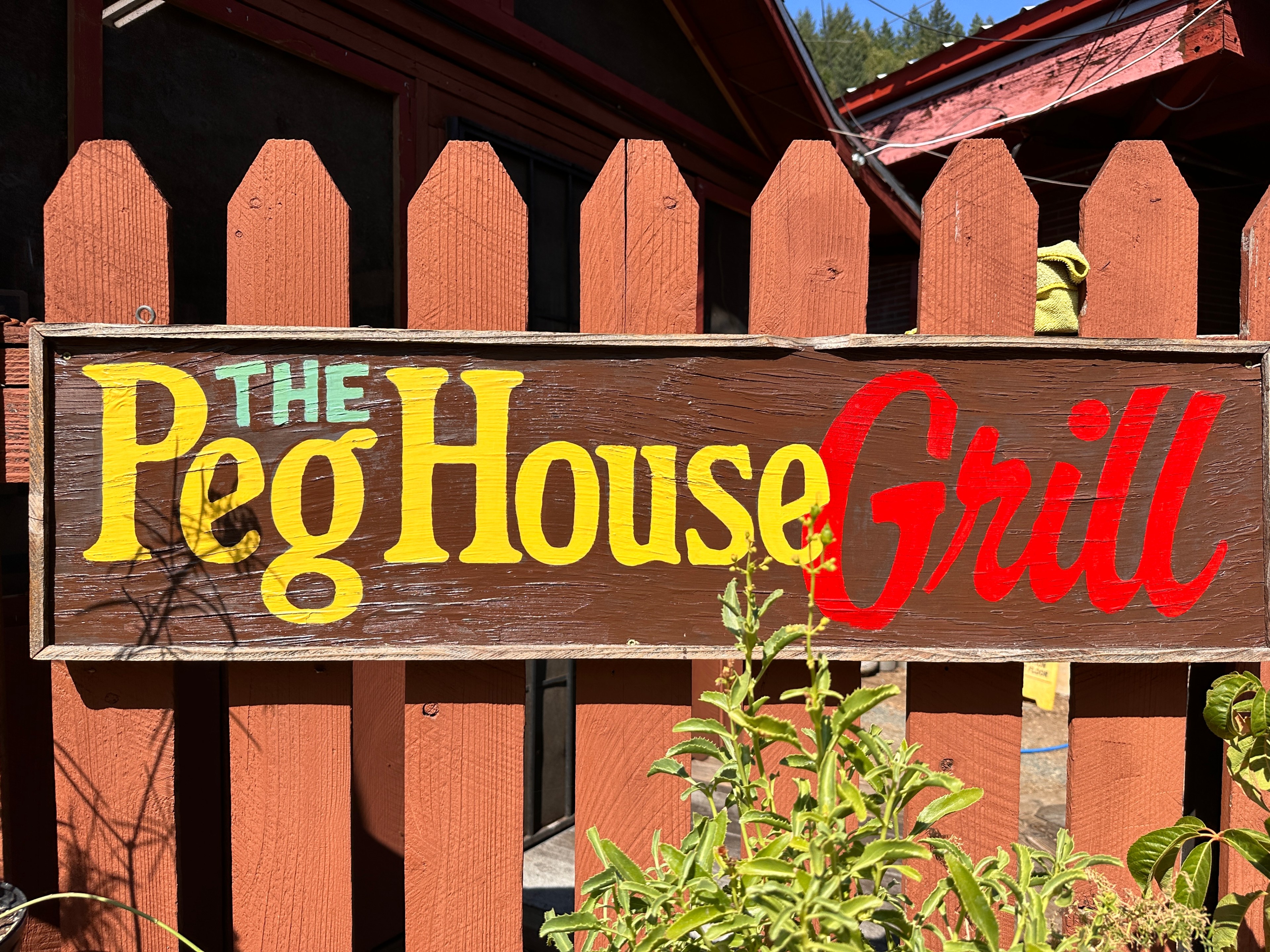 The image shows a wooden sign with &quot;The Peg House Grill&quot; painted in green, yellow, and red letters, mounted on a brown picket fence with some green plants in front.